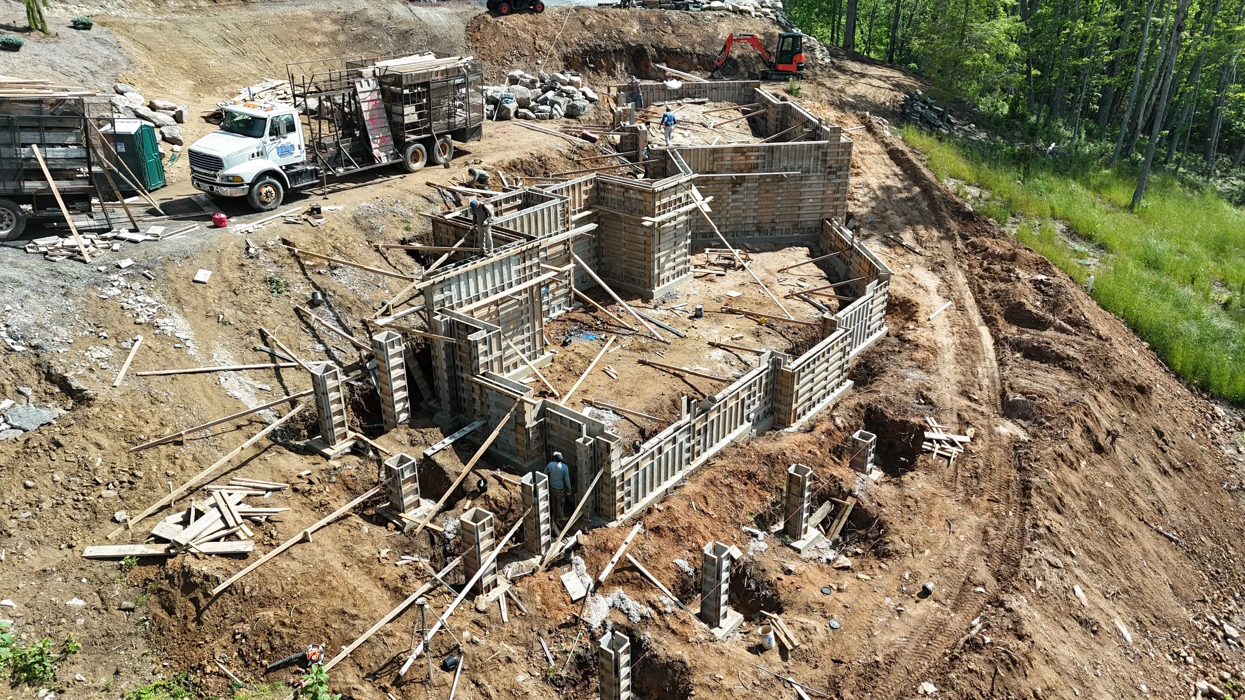 Construction site with concrete forms for a building foundation, workers, and construction vehicles amidst dirt and greenery.