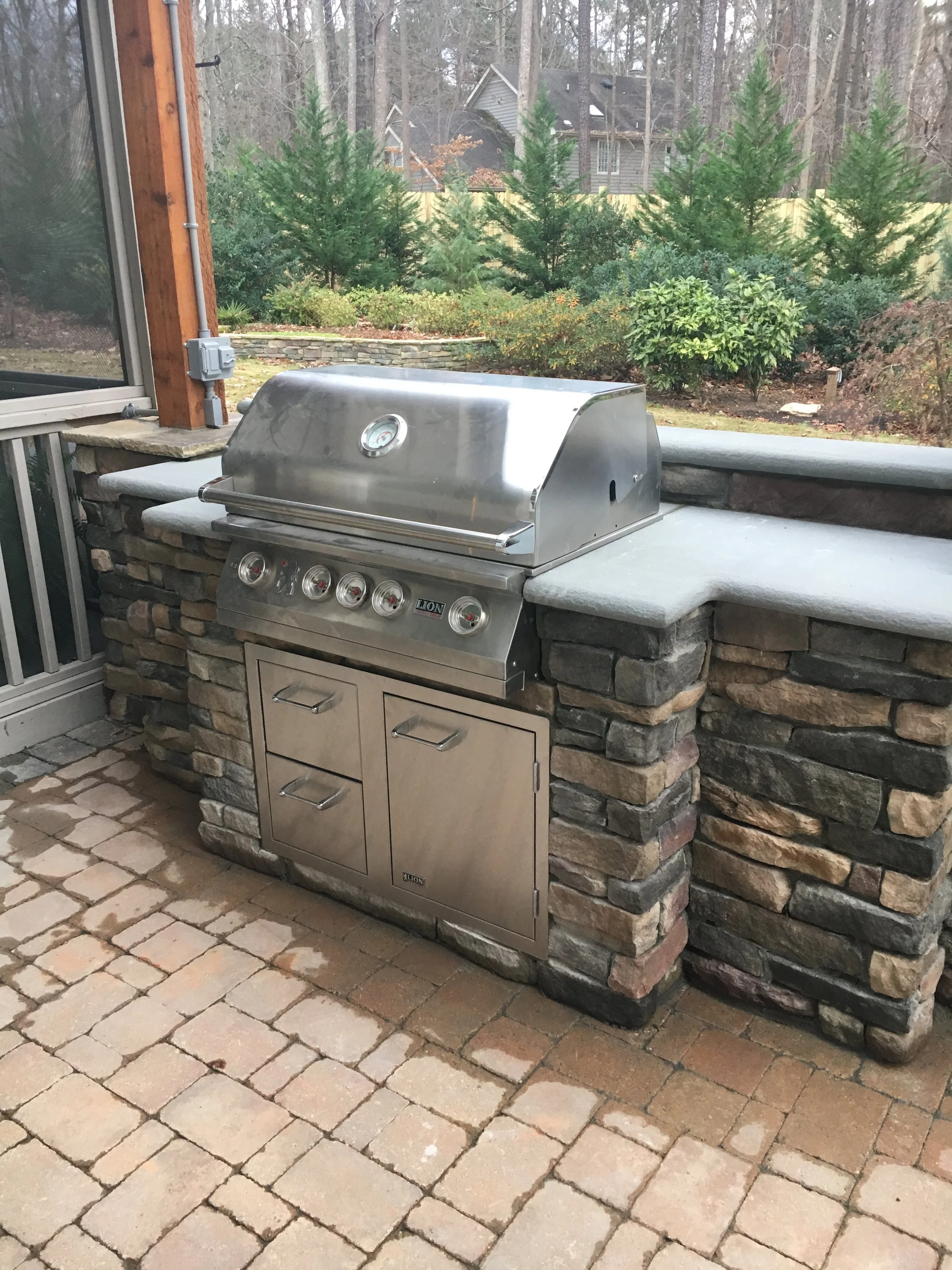A stainless steel outdoor gas grill built into a stone and concrete outdoor kitchen area with a brick patio floor and a wooded backyard with shrubs and trees.