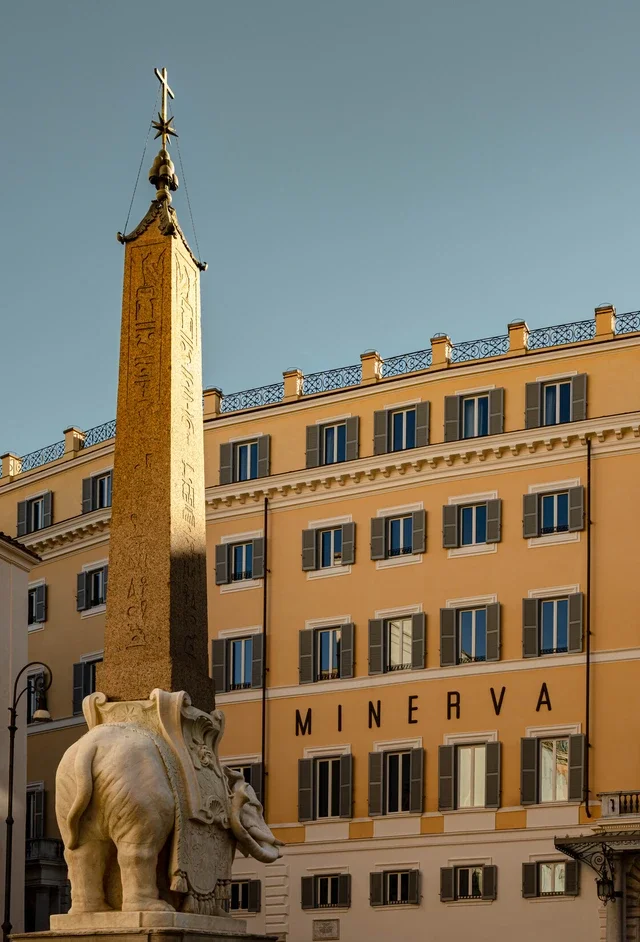 Ancient Egyptian obelisk with hieroglyphics on a pedestal featuring an elephant sculpture, located in an urban setting with a 19th-century style yellow building with windows and the sign 'MINERVA' in the background.