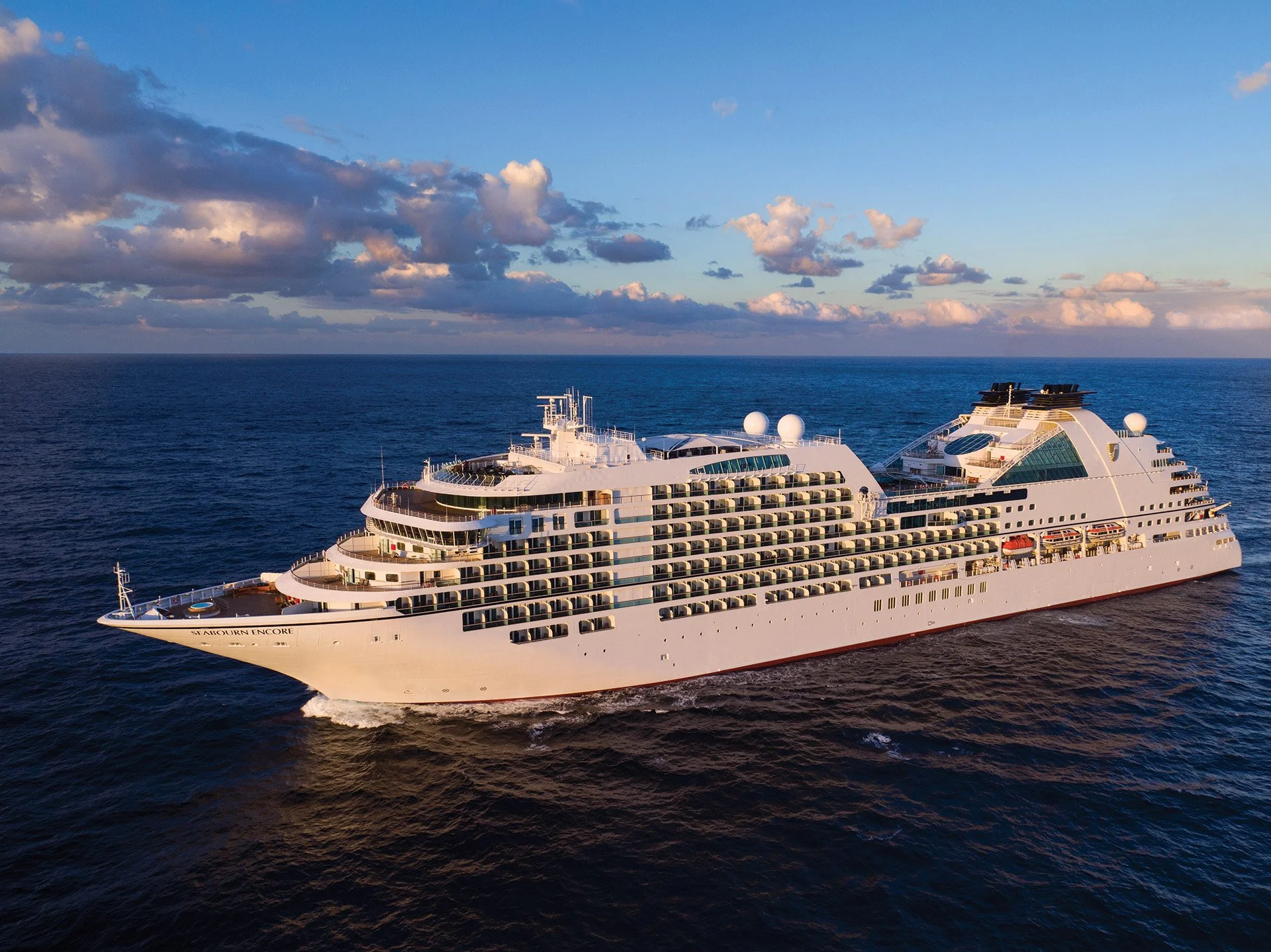 A large white cruise ship sailing on the open ocean under a partly cloudy sky at sunset.