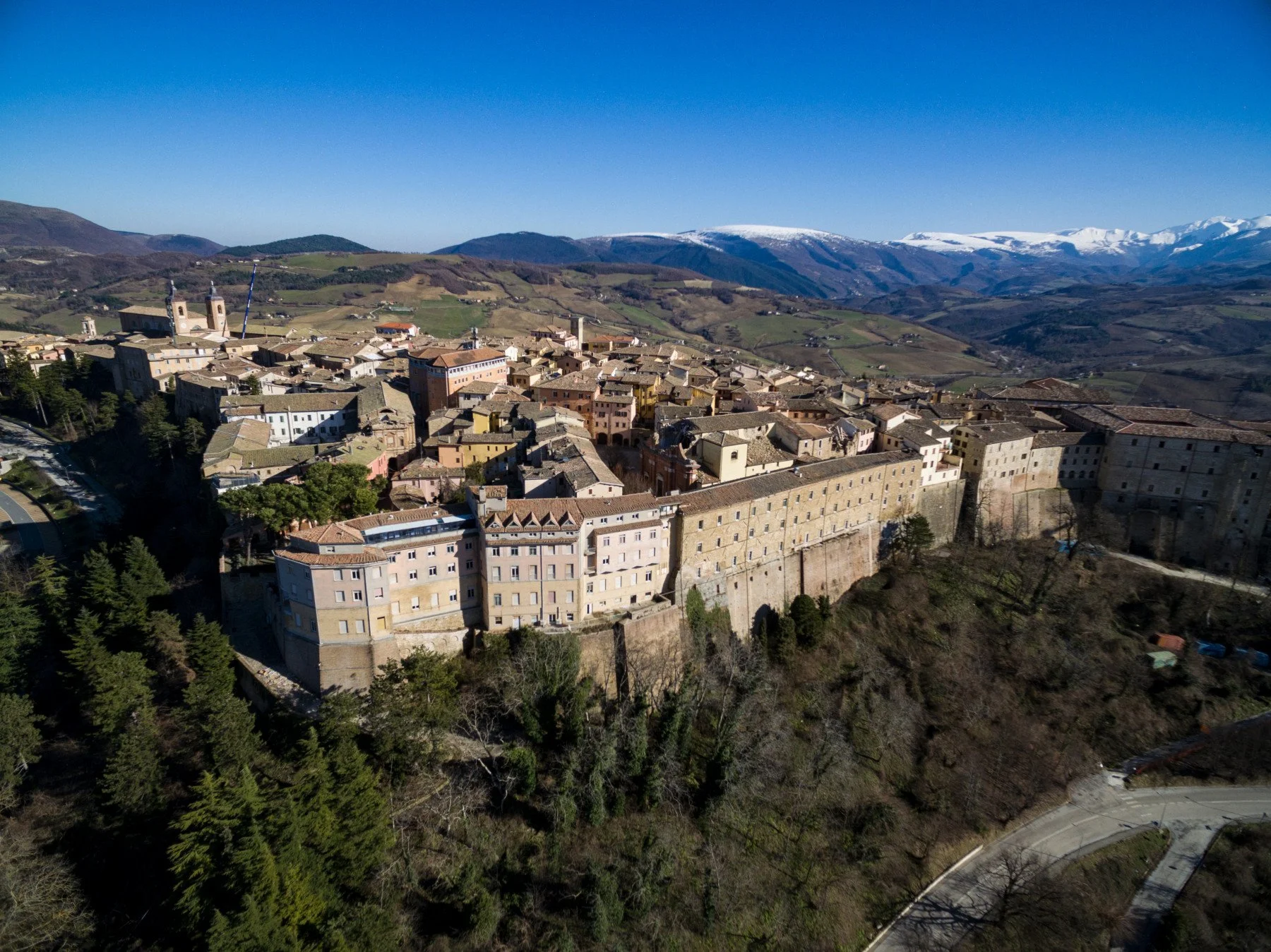 A panoramic view of a historic Italian hilltop town with medieval walls, surrounded by green hills and mountains in the background, under a clear blue sky.