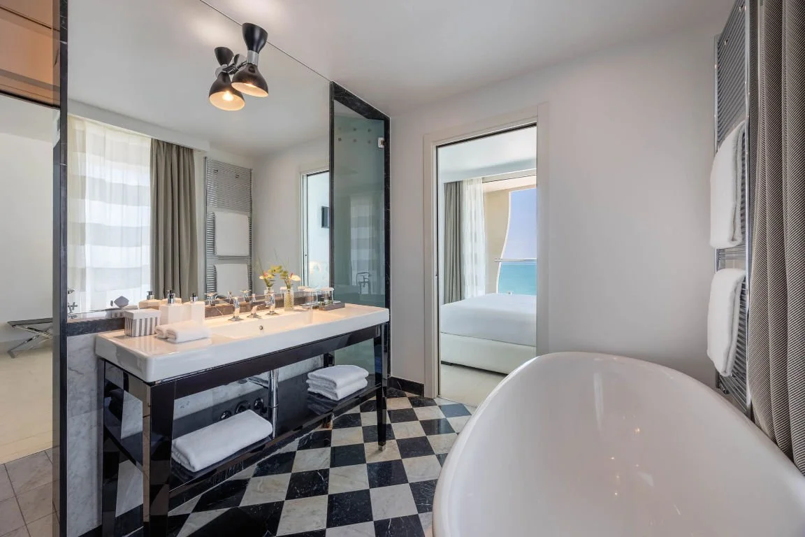 Modern bathroom with a black and white checkered floor, a white bathtub, a black-framed mirror, a double sink with toiletries, a window with curtains, and a door leading to a bedroom with a bed and ocean view.