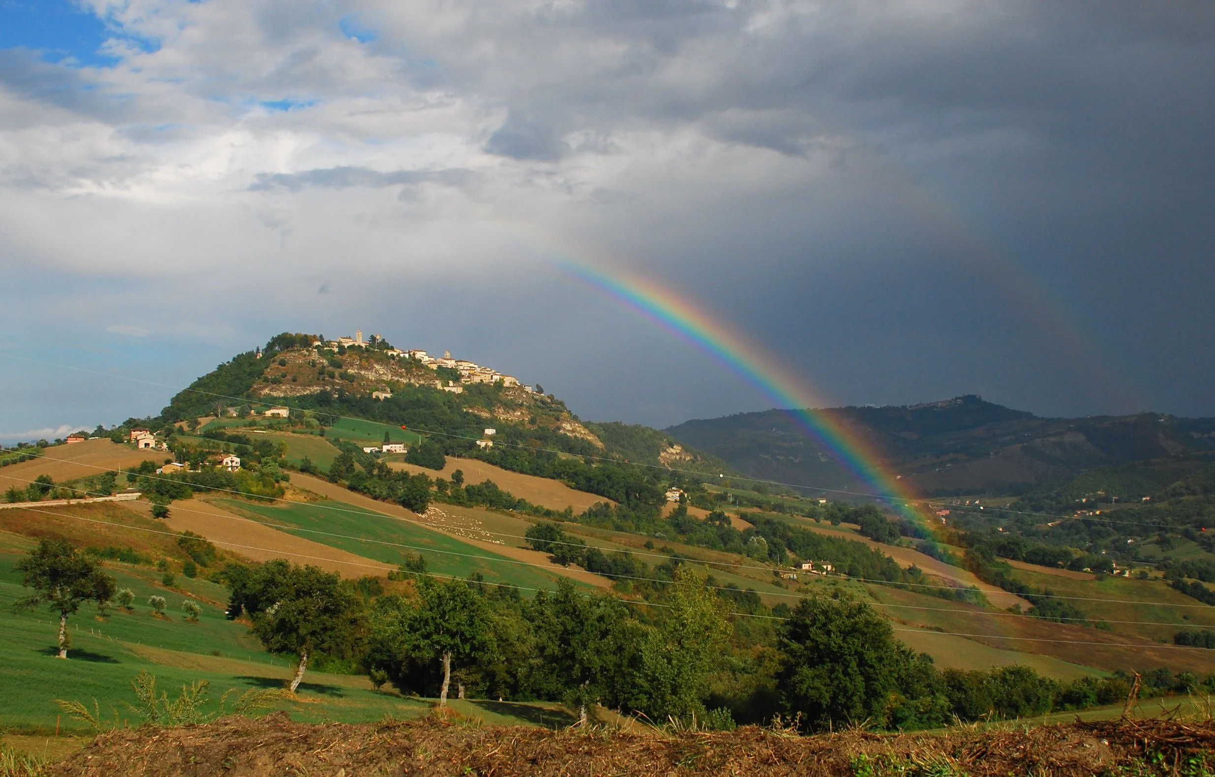 A rainbow arching over a green hillside landscape with houses and trees, under a cloudy sky.