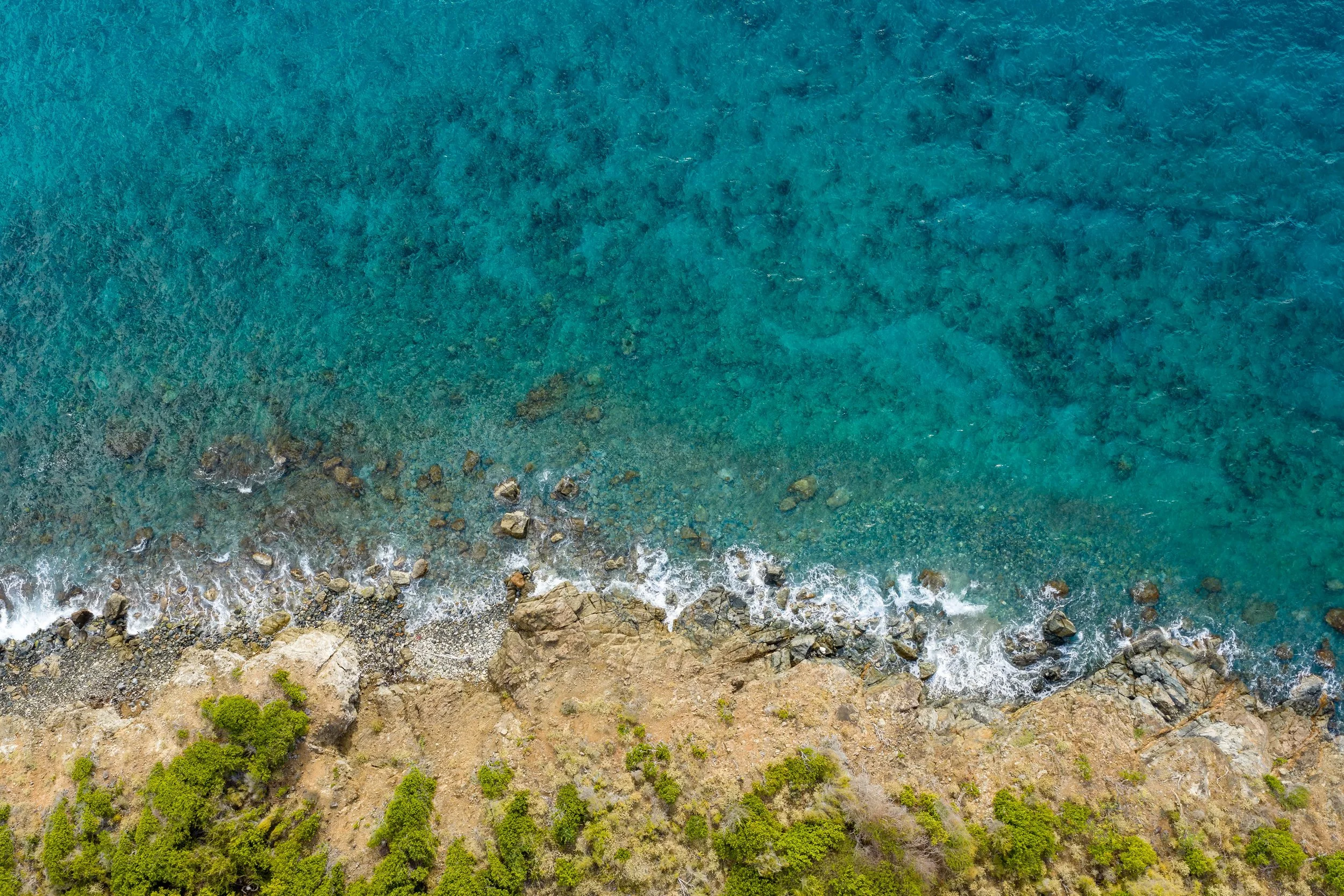 Aerial view of a rocky shoreline with clear blue-green ocean water, small waves, and green vegetation at the coast.