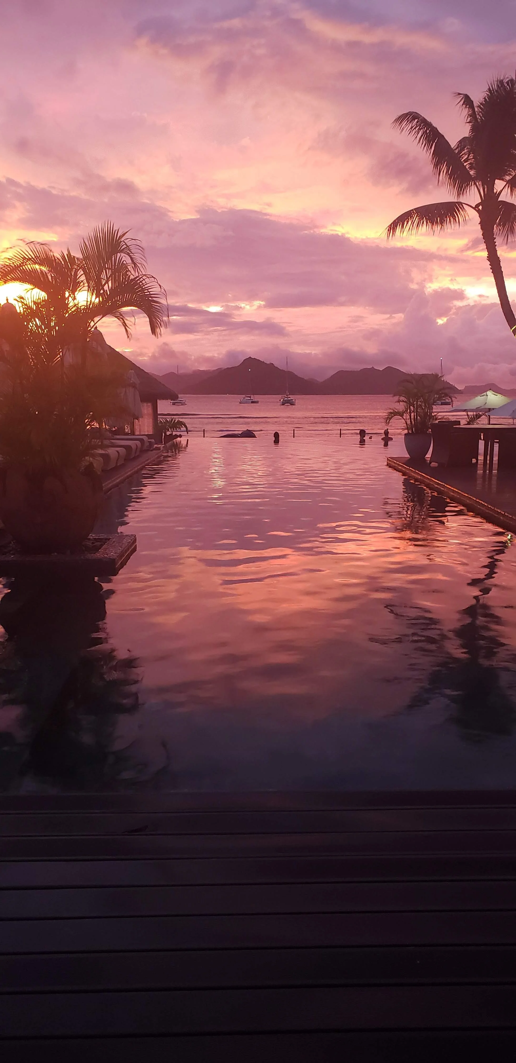 Sunset over a tropical ocean view with palm trees, mountains, sailboats, and a pool reflecting the colorful sky.