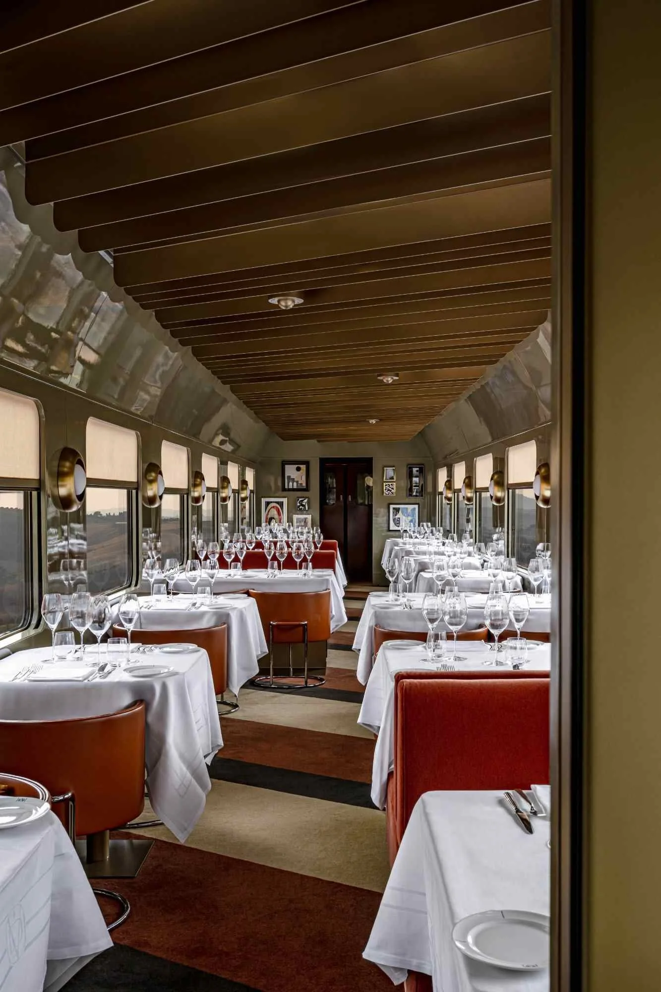 The image shows the interior of a restaurant dining area with neatly set tables covered with white tablecloths, wine glasses, and silverware. The room has large windows along the sides, framed by gold-colored light fixtures, and the ceiling features wooden slats.