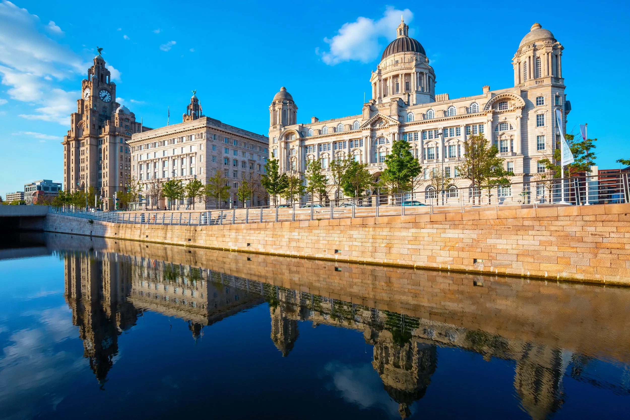 The Liverpool waterfront in England with historic buildings reflected in the calm river under a partly cloudy blue sky.