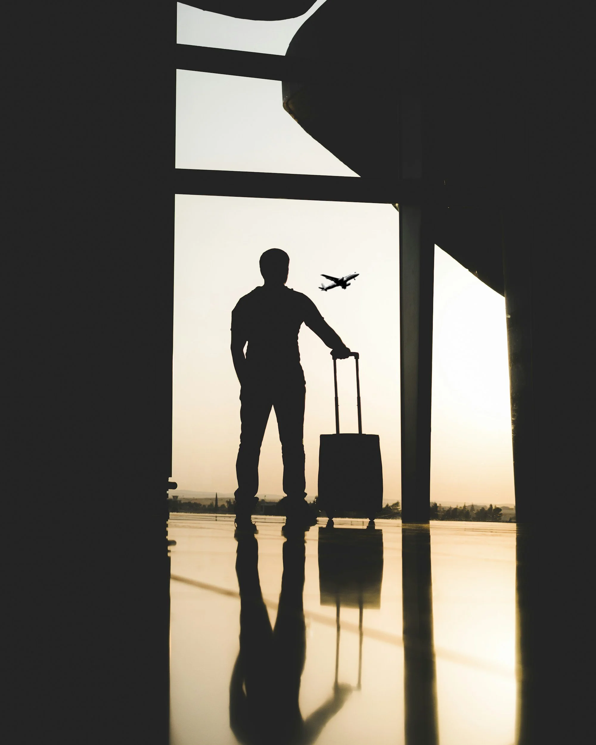 Silhouette of a man carrying a suitcase, standing at an airport terminal window, with an airplane flying in the sky outside during sunset.