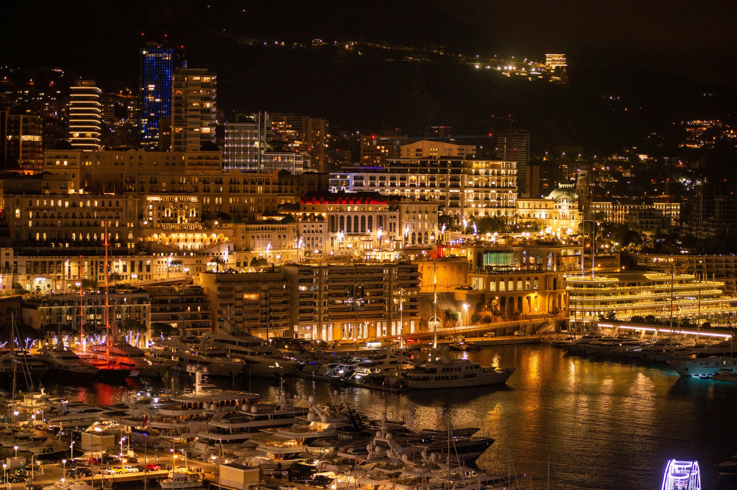 Nighttime cityscape with illuminated buildings, yachts, and boats docked at a marina with a hill in the background, lit with various lights.