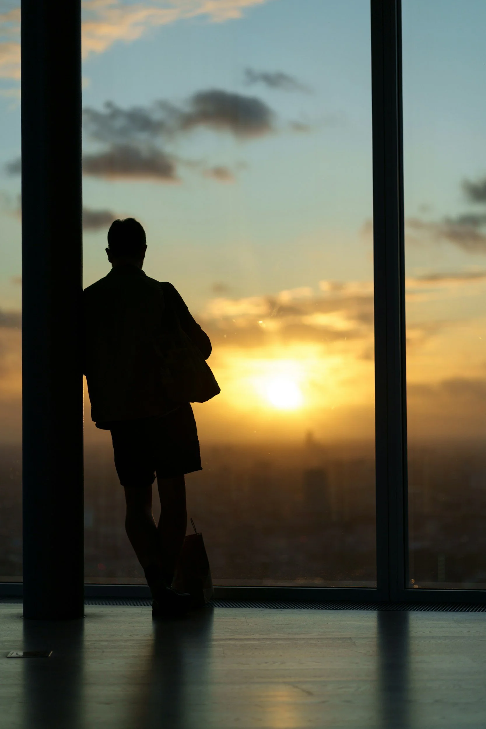 A person standing indoors near a large window, looking outside at a sunset sky with clouds.
