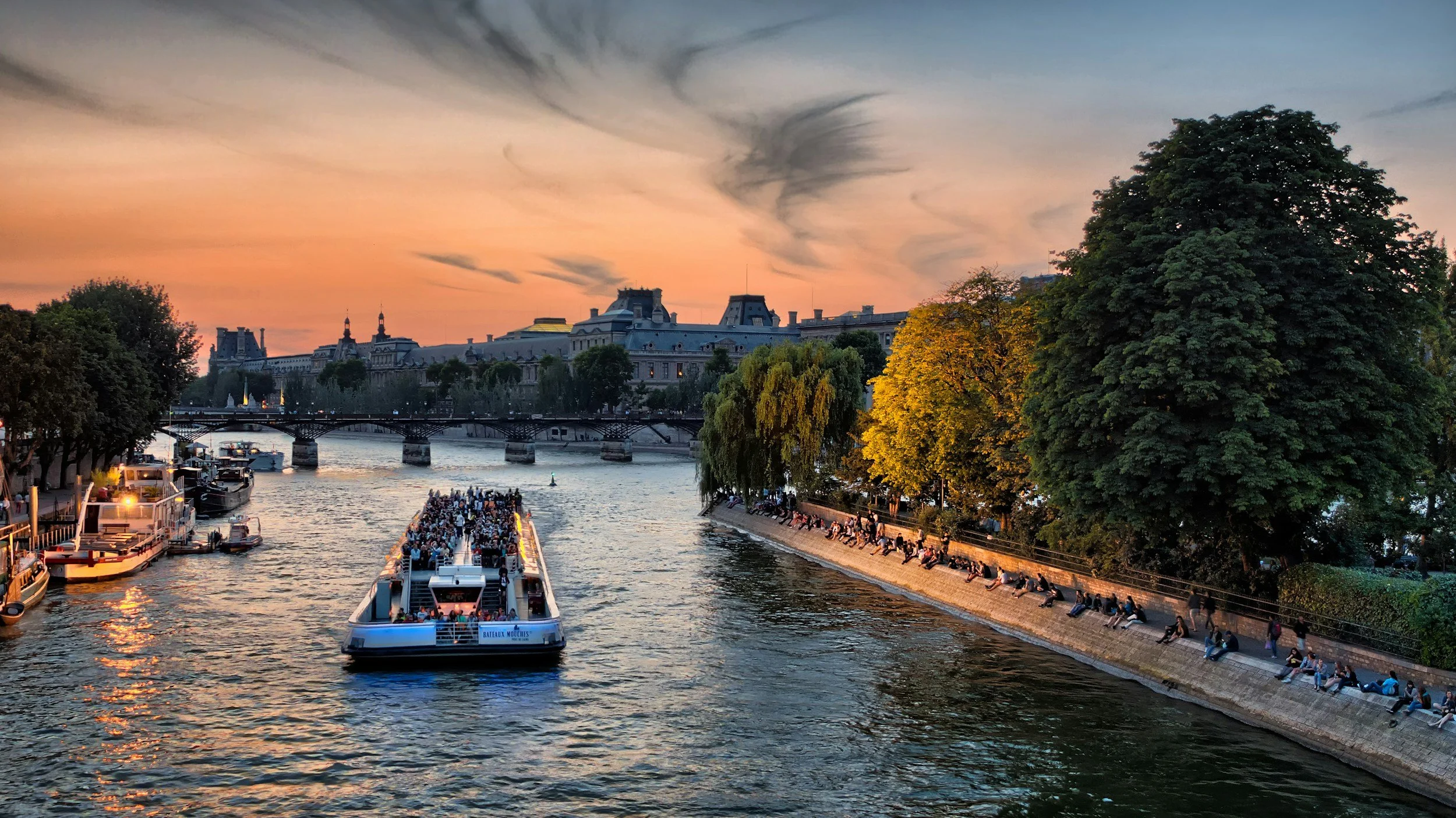 A sunset view of the Seine River in Paris with boats and people along the riverbank, trees, and historic buildings in the background.