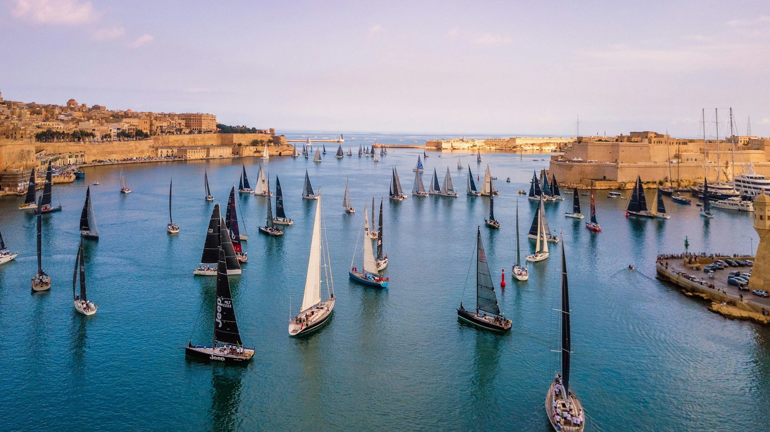 Marina filled with sailboats and yachts, with historic city walls and buildings in the background, set against a calm sea and partly cloudy sky.
