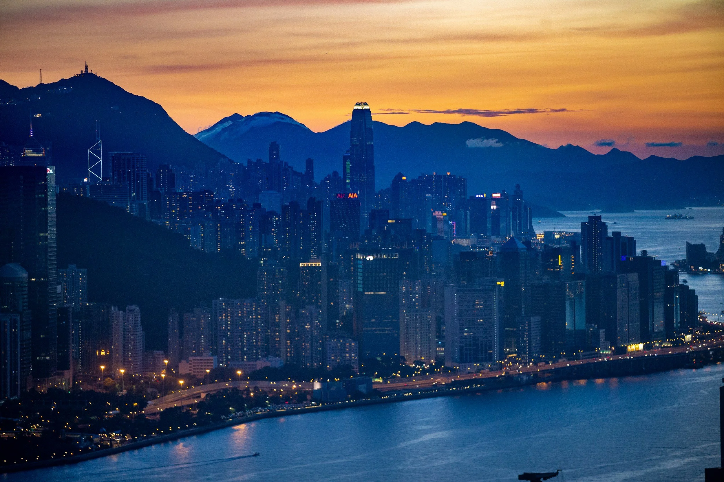 A city skyline at dusk with tall buildings, mountains in the background, and a body of water in the foreground.