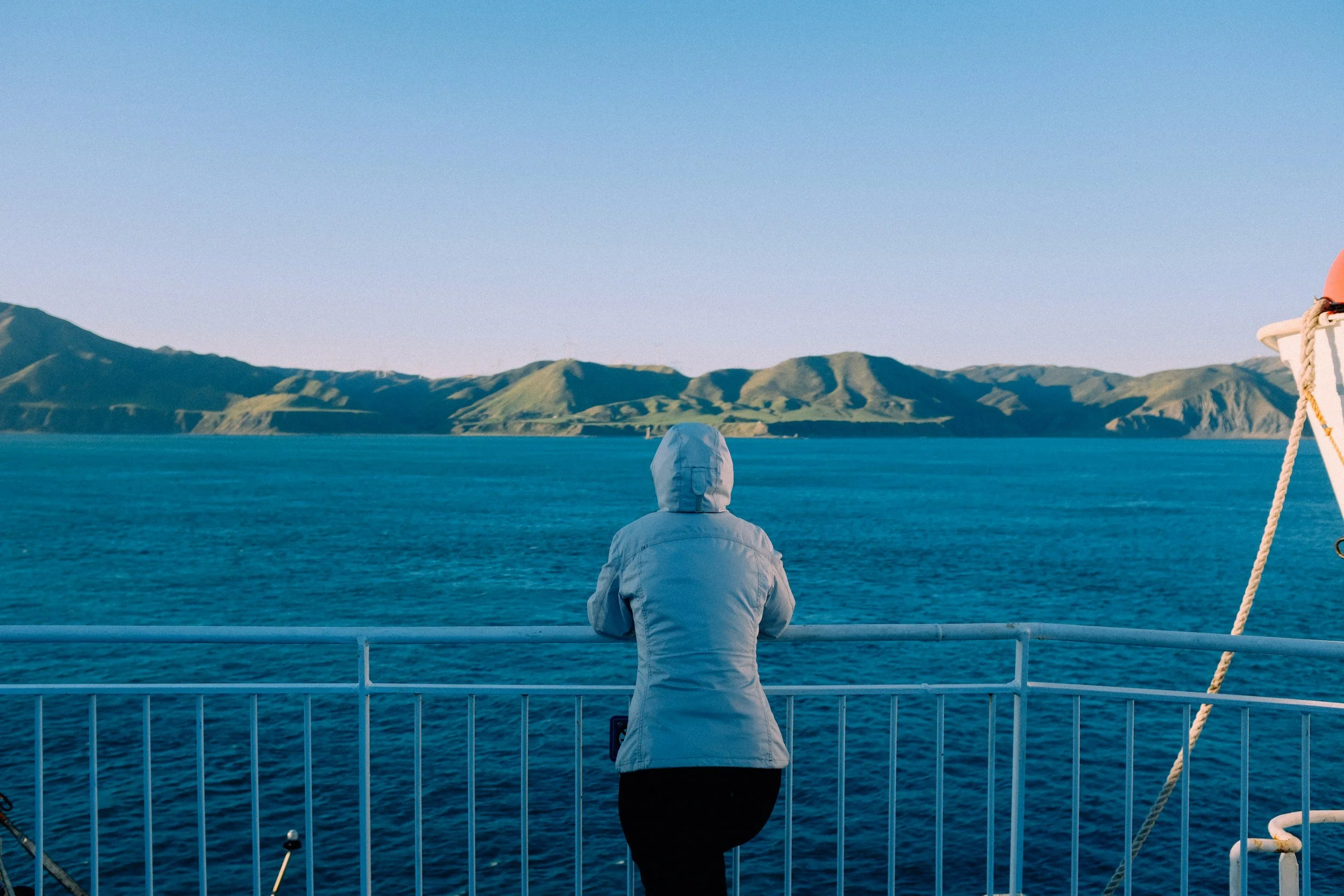 Person in a white jacket and hood facing away, standing on the deck of a boat, looking toward distant mountains and blue water.