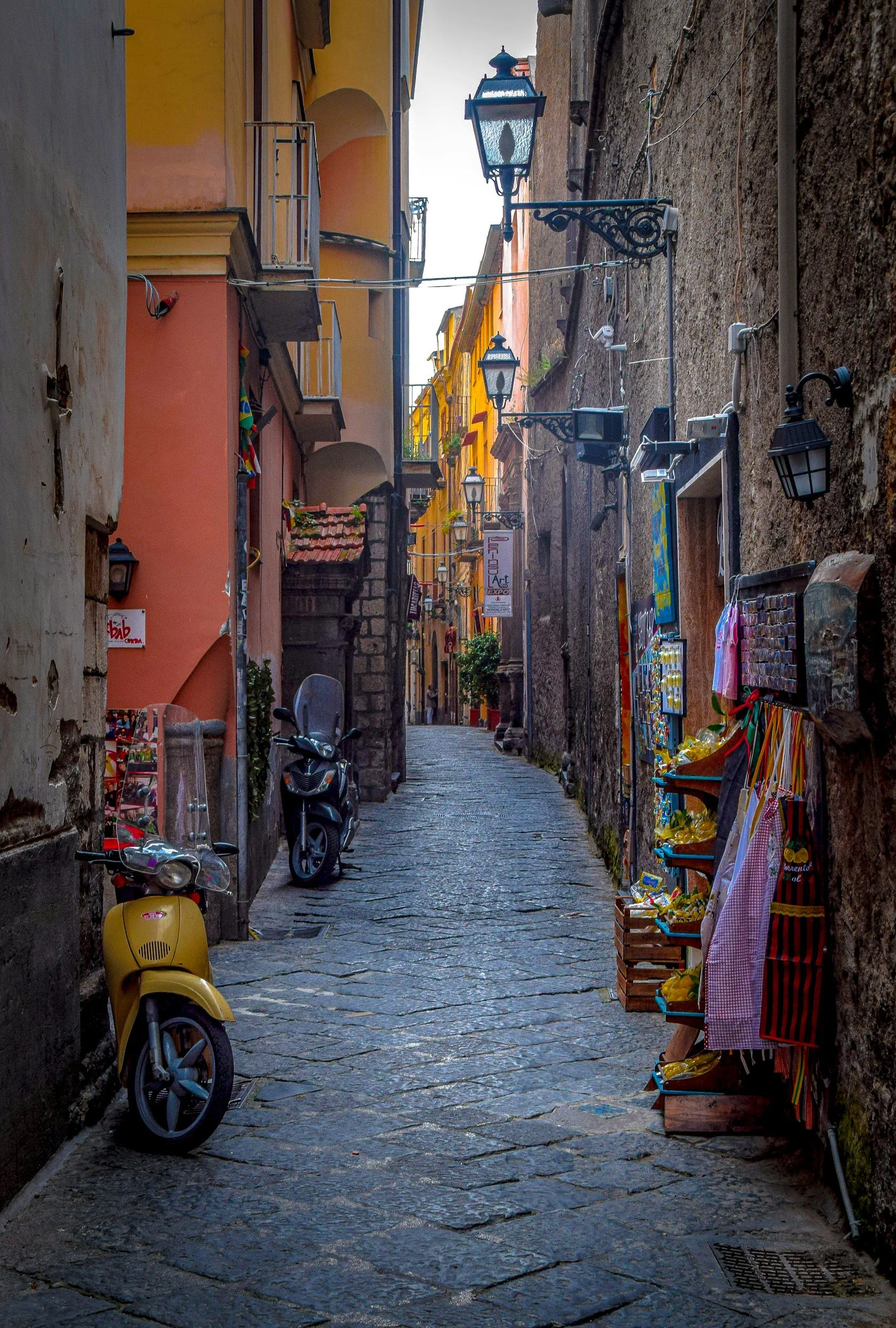 A narrow cobblestone street lined with colorful buildings featuring small balconies and hanging lanterns. There are parked scooters and a small outdoor market with clothes and souvenirs on display along the sidewalk.