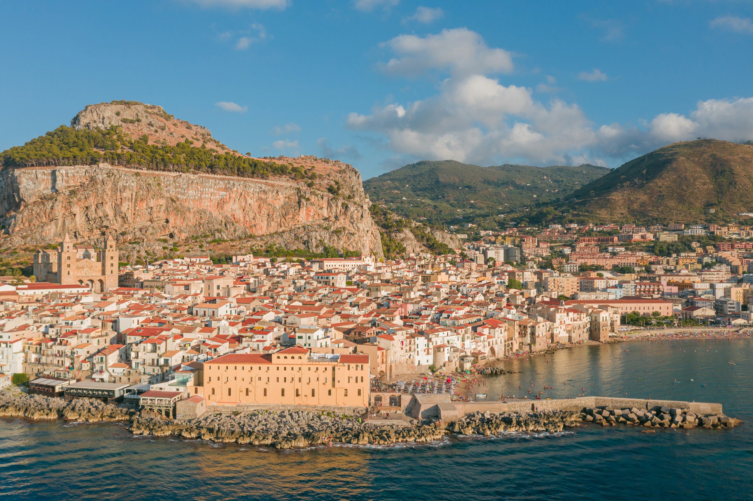 A coastal town with tightly packed buildings along the shoreline, a church with twin towers, a mountain with greenery and rock formations, and a clear blue sky with some clouds.