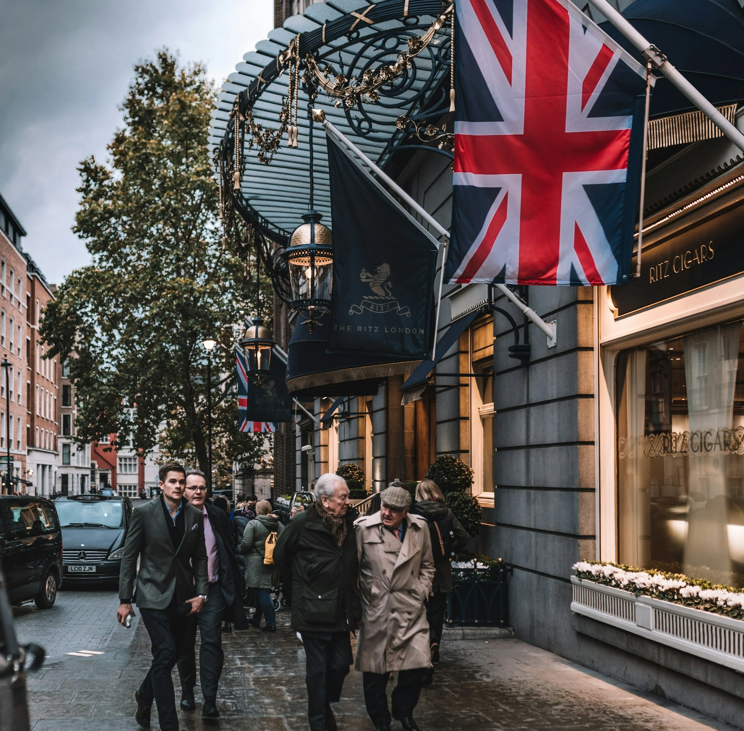 People walking on a city sidewalk in front of shops and flags, including a prominent British flag, on a cloudy day.