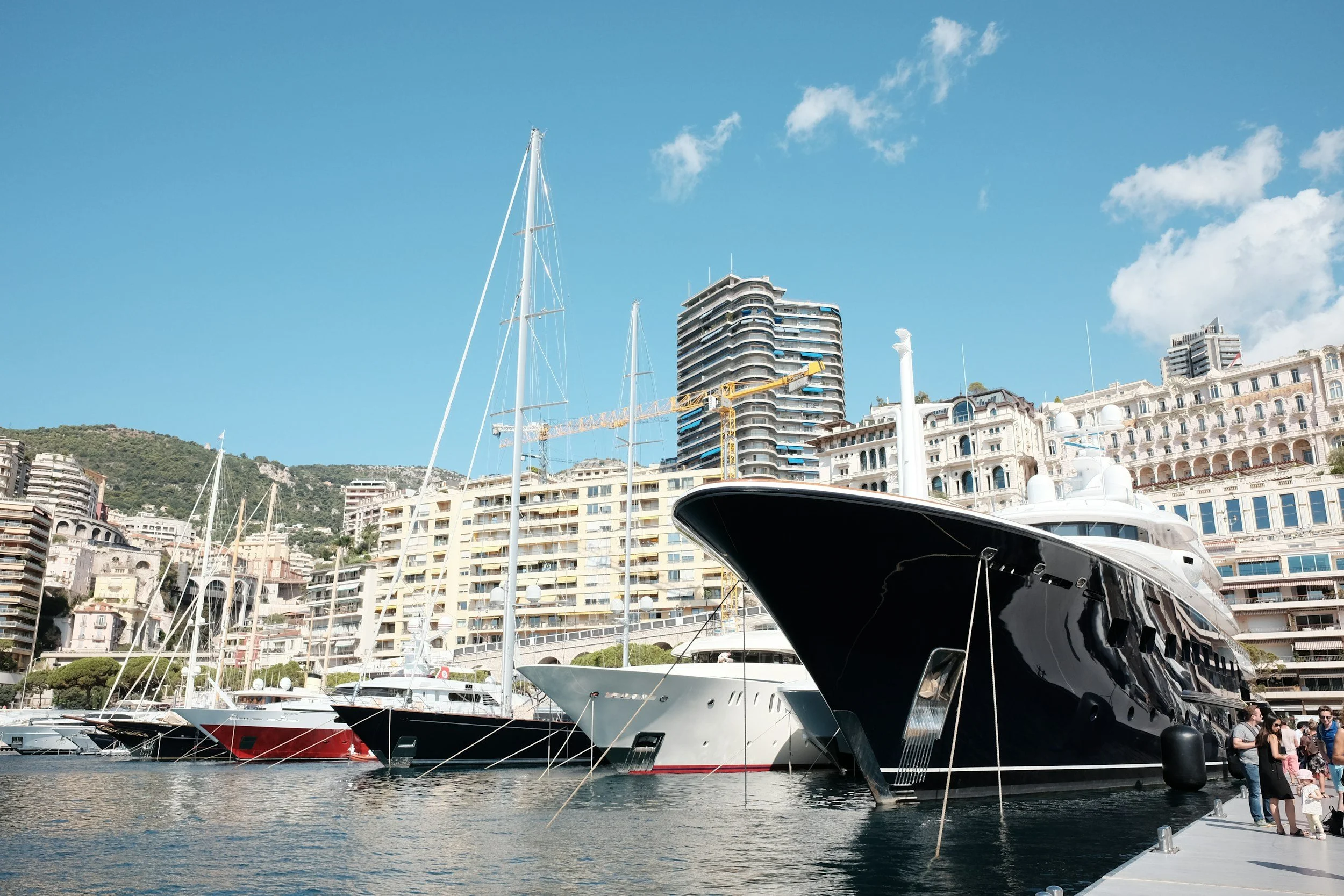 Luxury yachts docked at a marina with high-rise buildings and a hillside in the background under a partly cloudy blue sky.