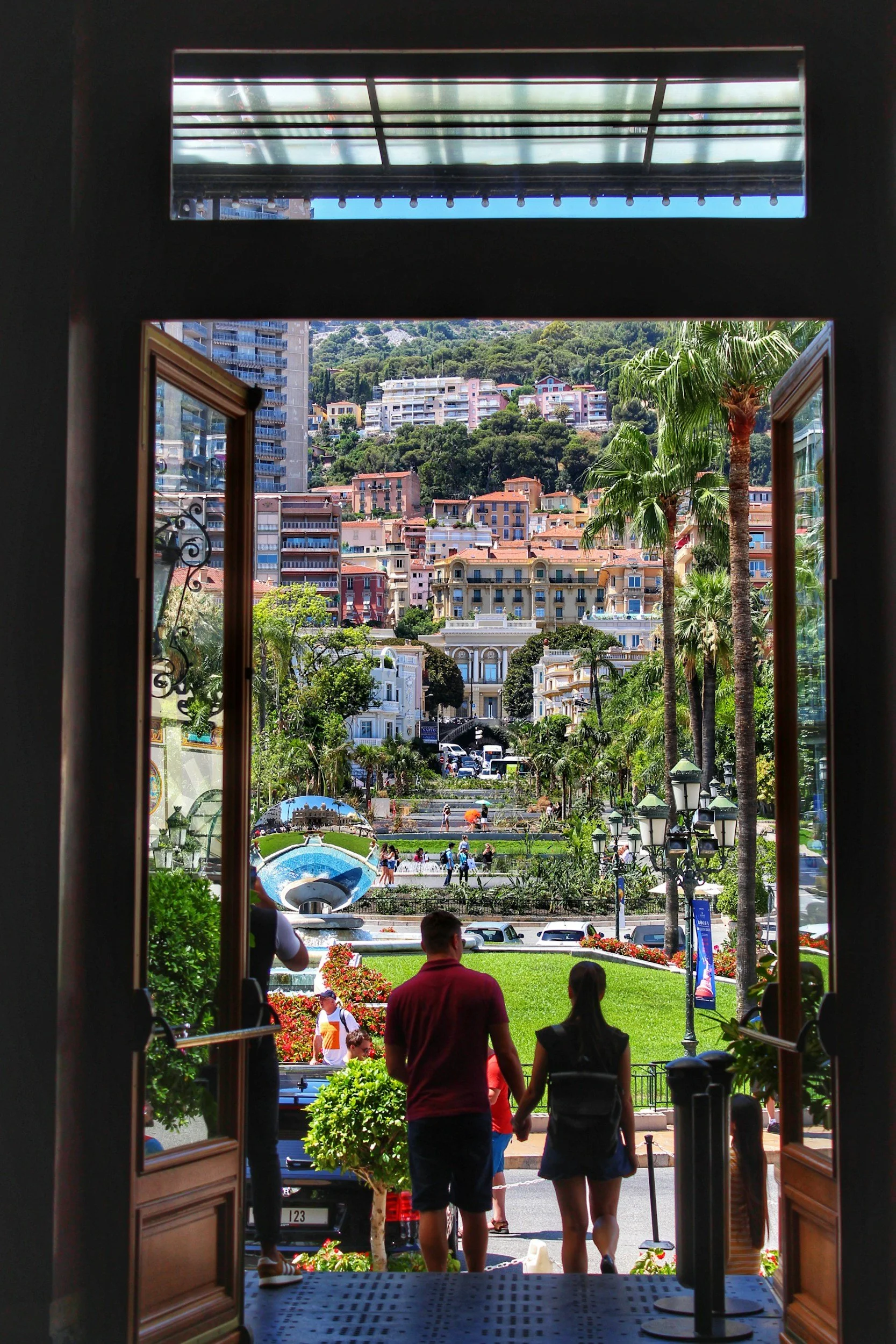 People walking out of a building into a colorful cityscape with palm trees, multi-story buildings, and a park with a fountain, sunny weather.