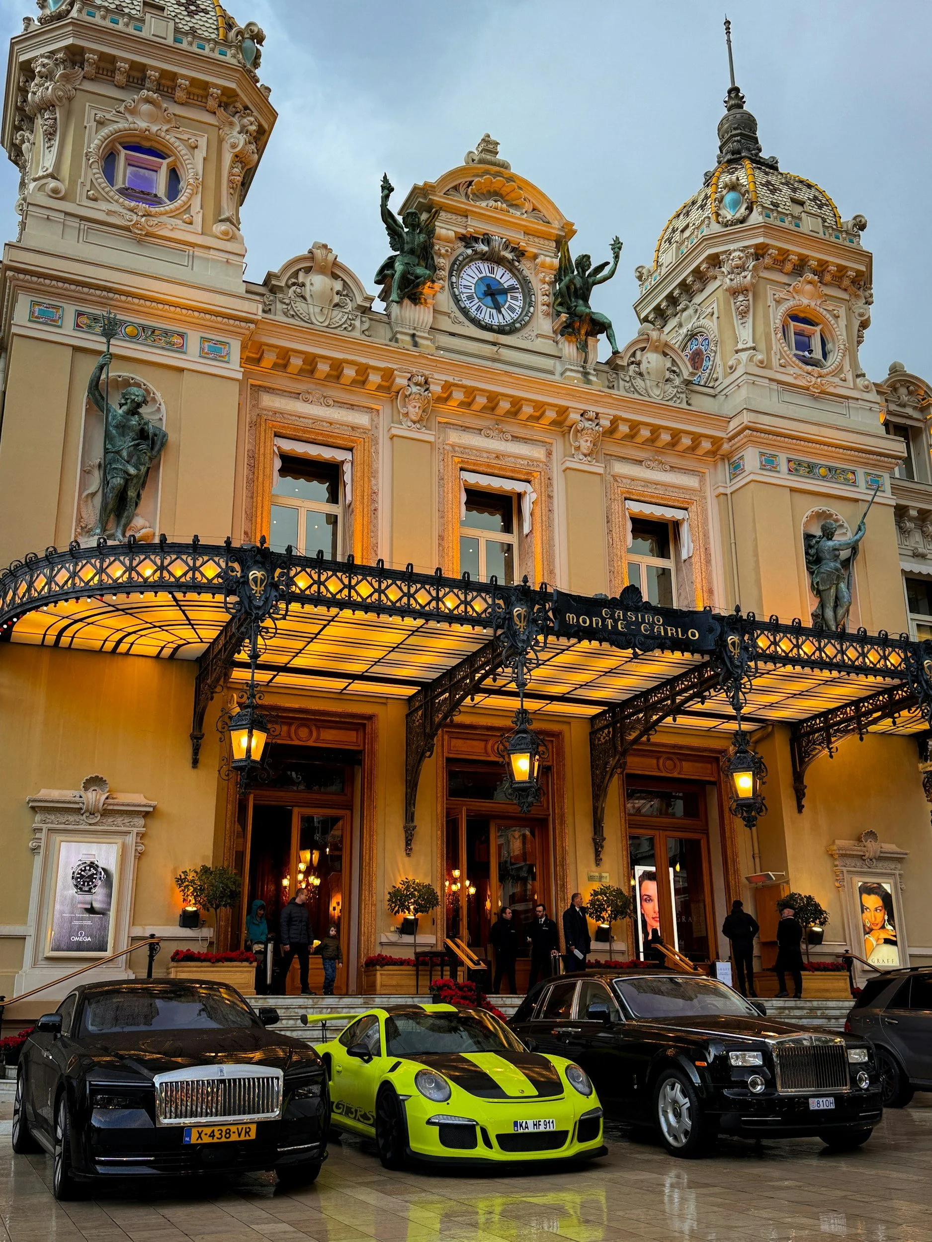Luxury casino hotel building with ornate architecture, statues, and a clock on the facade. Several luxury cars are parked in front of the entrance on a wet pavement.