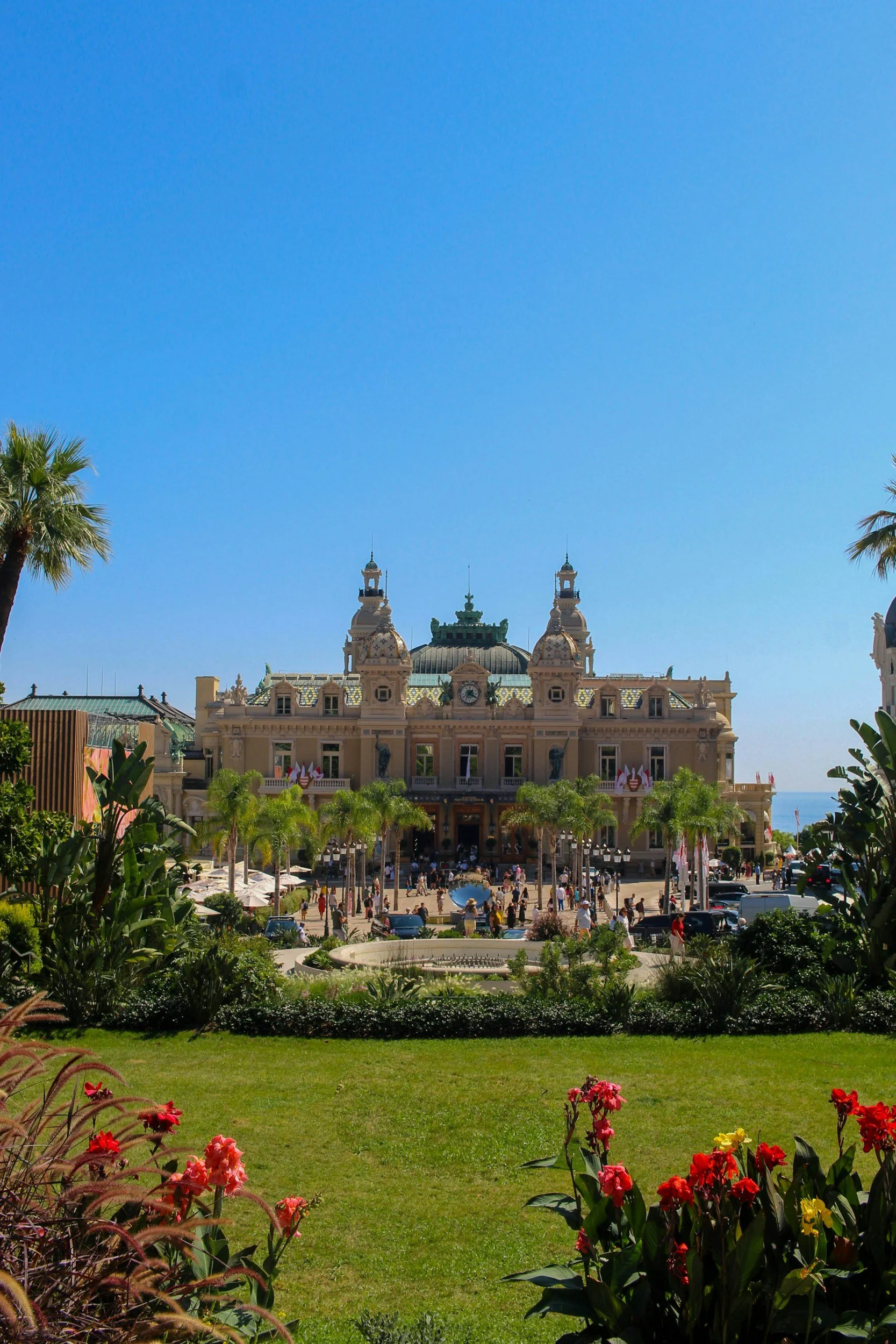 A large, ornate building with a green dome and towers, surrounded by palm trees and lush gardens, with a crowd of people and cars in front.
