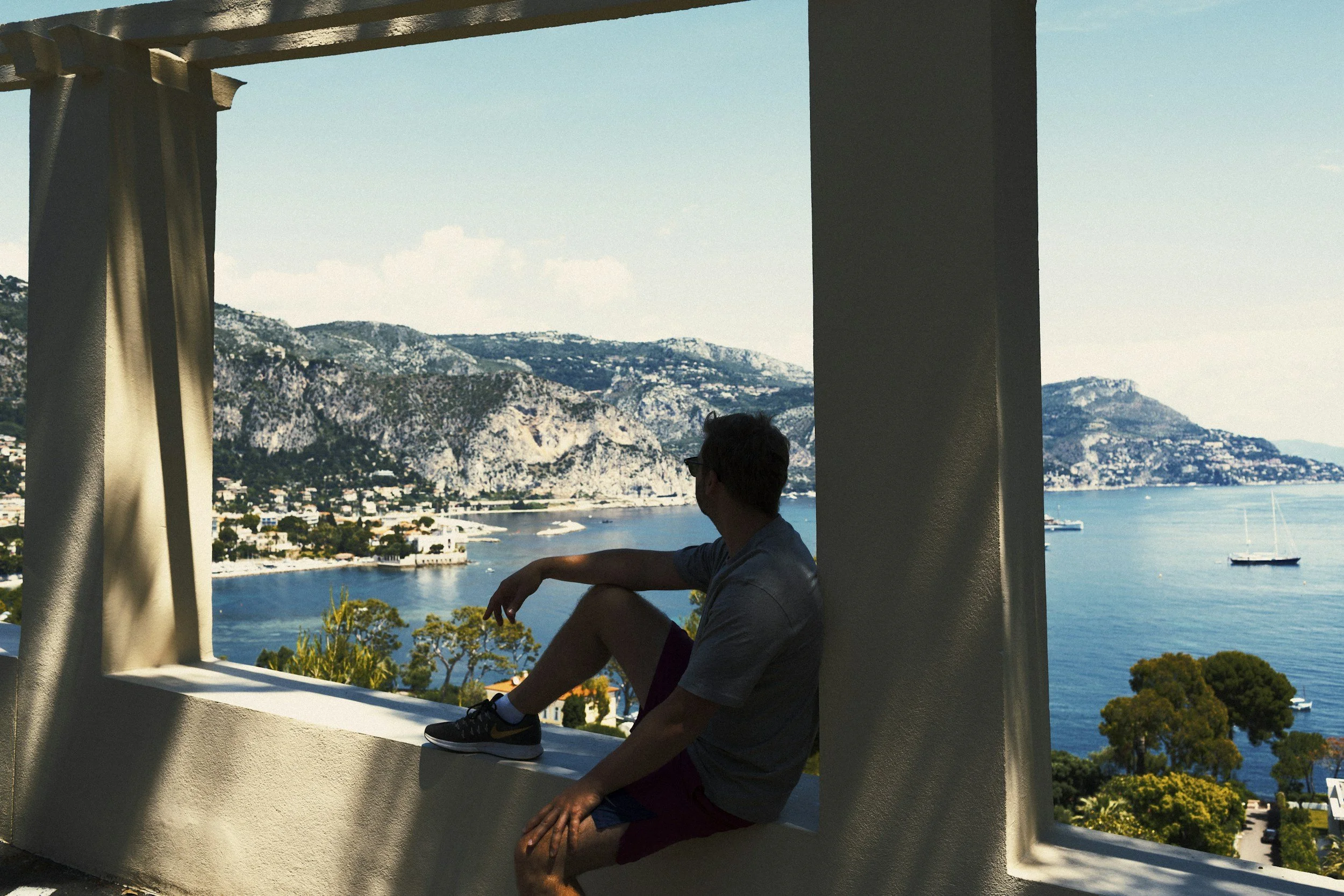 A man sitting on a ledge with a scenic view of a bay, boats, and mountains in the background, partly shaded by a structure with columns and curtains.