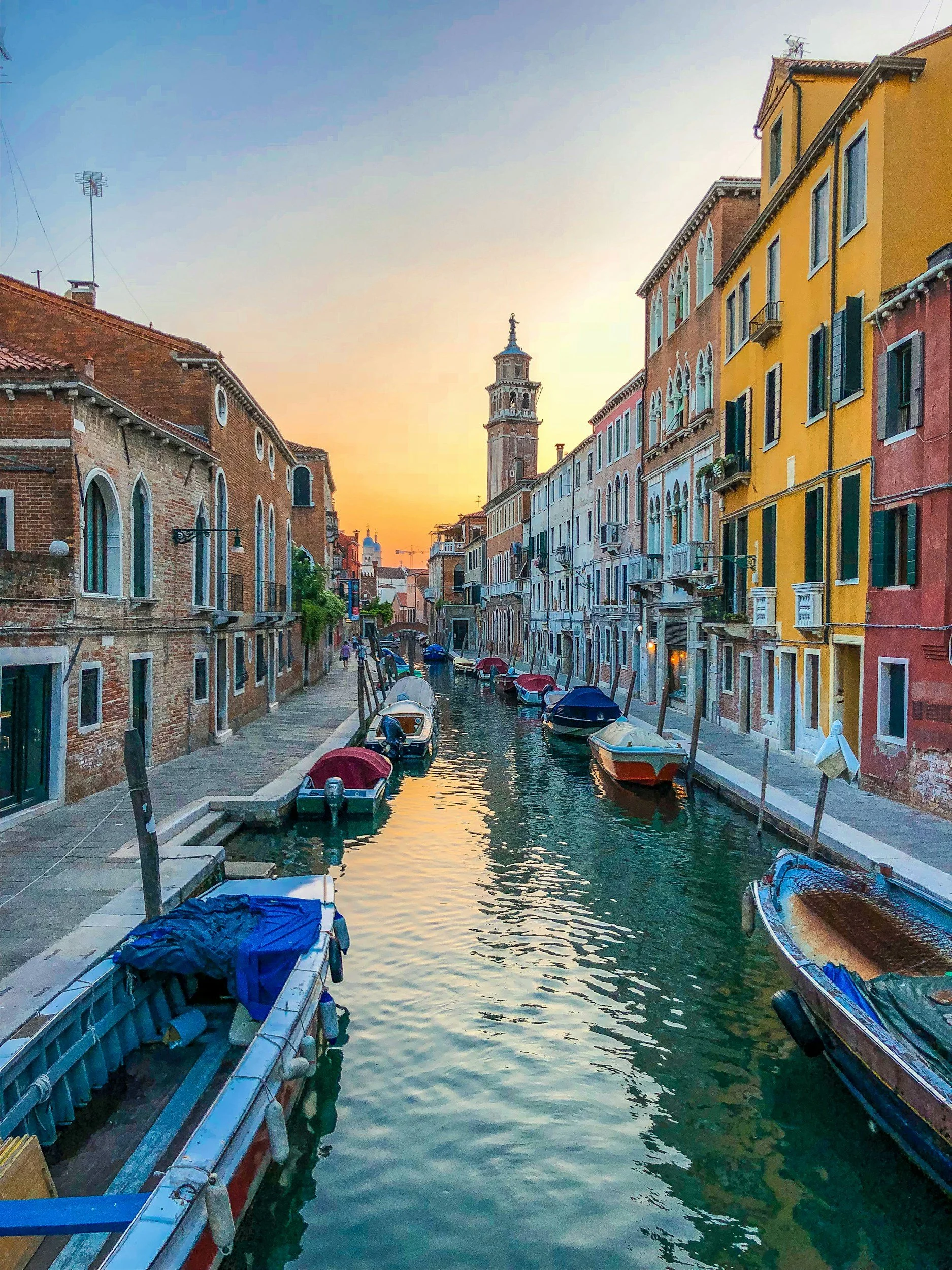 Colorful buildings and boats along a canal during sunset, with a clock tower in the distance.
