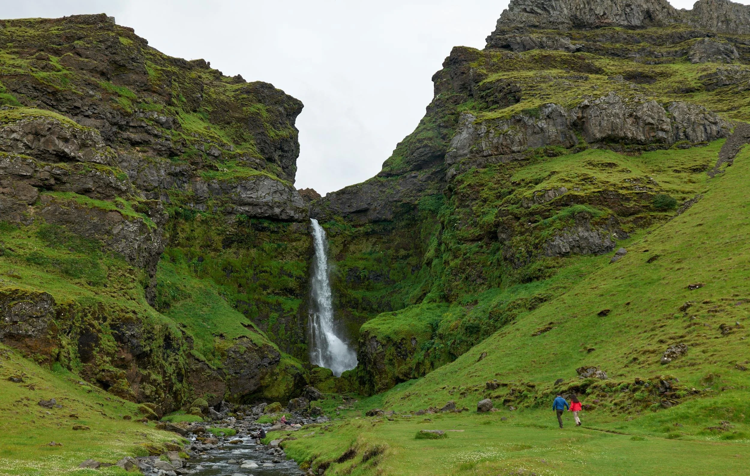 Two people walking along a grassy path near a small river, with a waterfall cascading down moss-covered cliffs in the background.