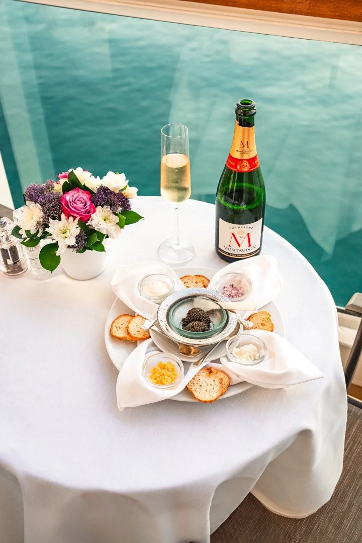 A table set for a special occasion with a flower arrangement, a glass of champagne, a bottle of champagne, and a tray of assorted caviar with bread and garnishes, near a window overlooking a body of water.