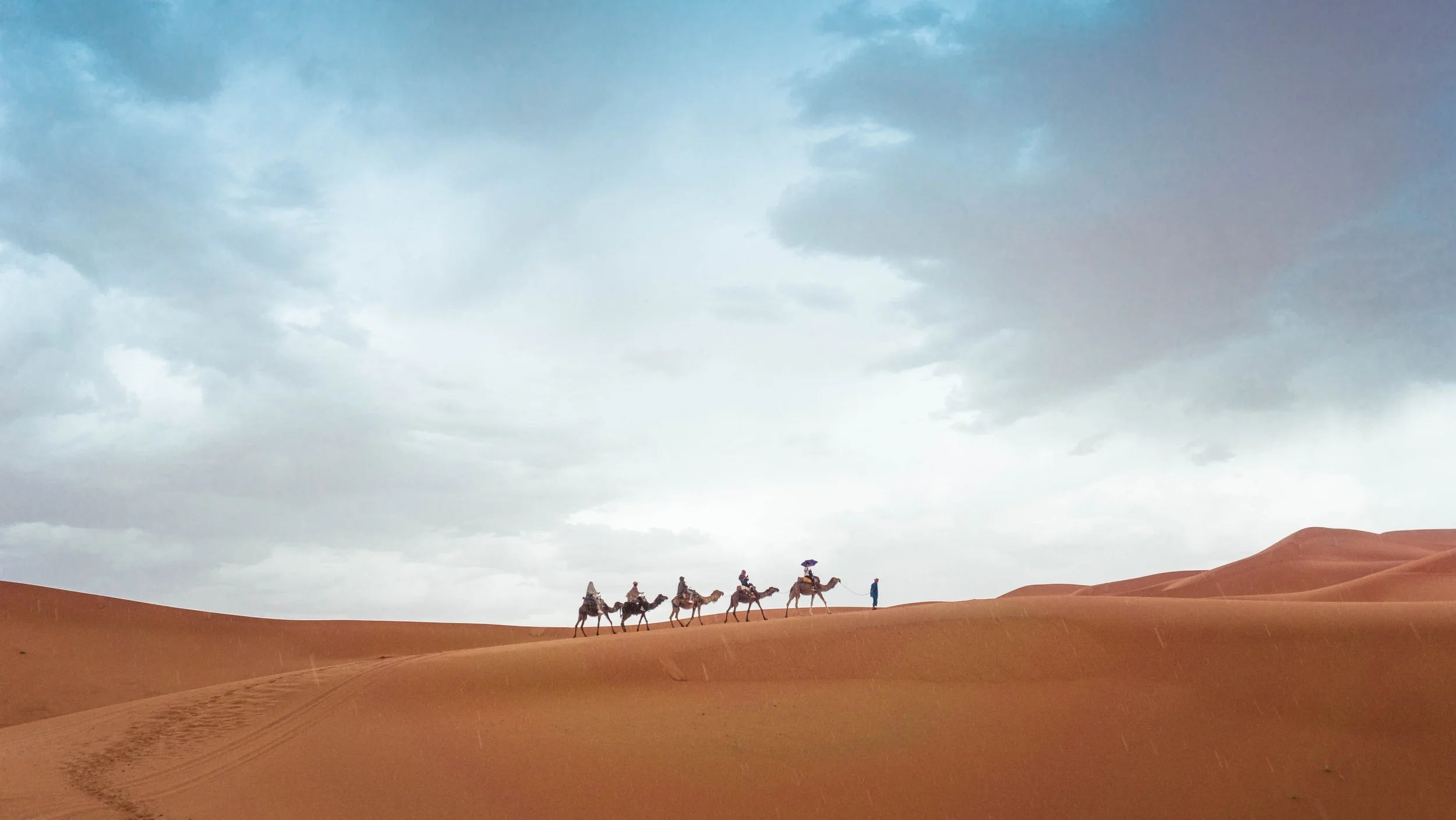 A group of people riding camels across the desert with a cloudy sky overhead.