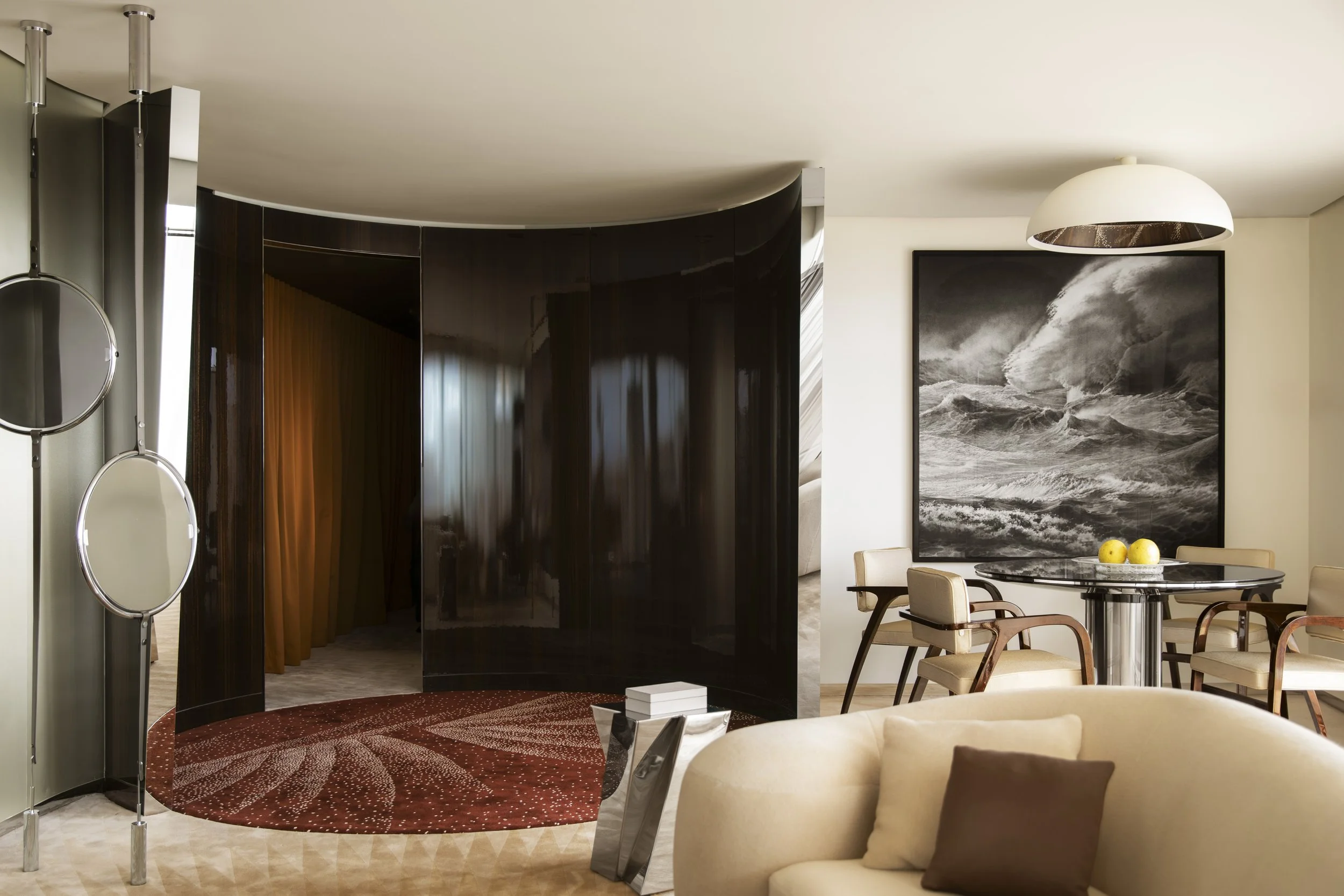 Living room with a round black and white painting of ocean waves, a dining table with beige chairs, a beige couch with brown pillows, a dark curved room divider, and a round red rug.