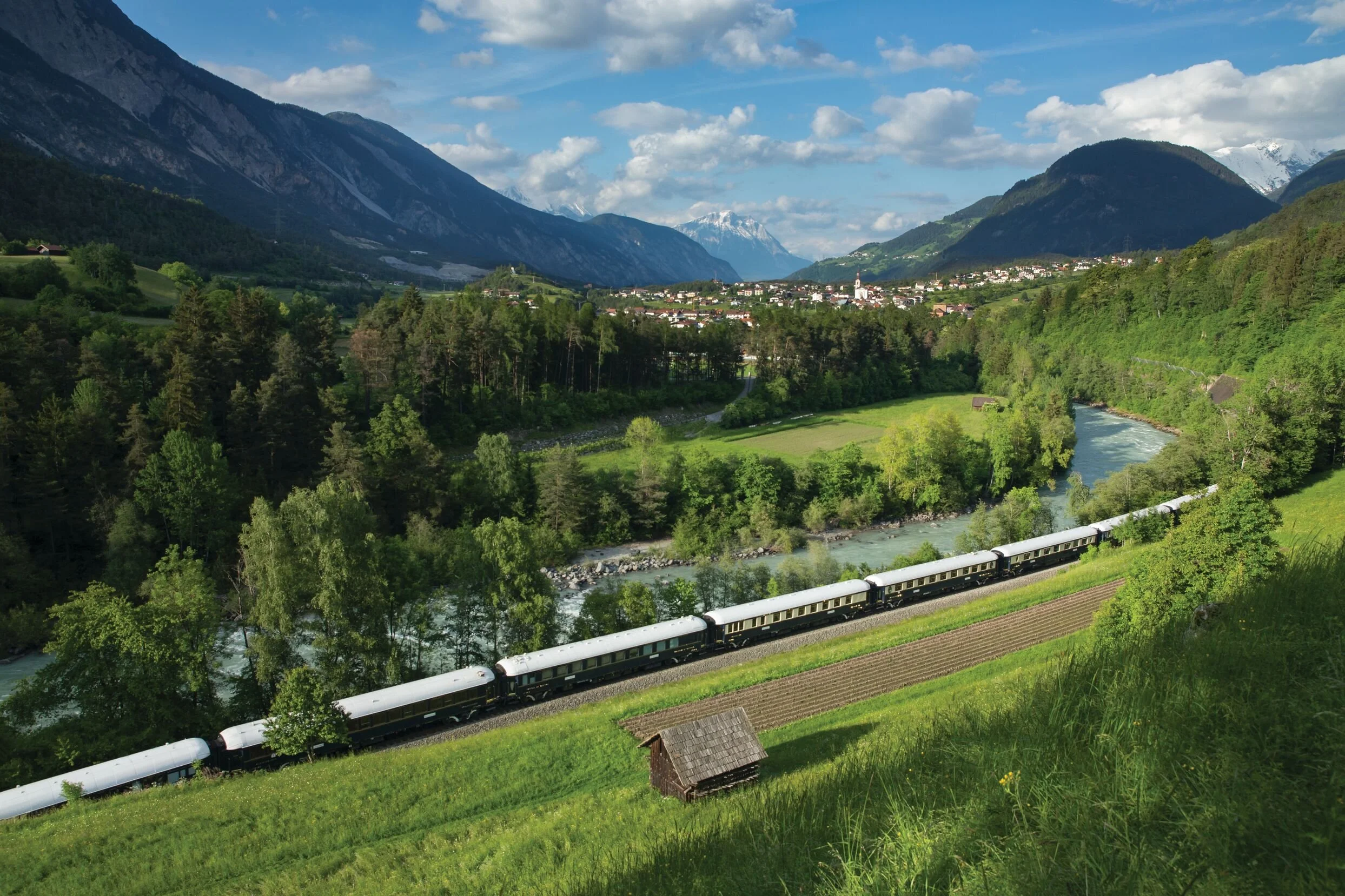 A scenic landscape of a green valley with a river running through it, surrounded by mountains with snow-capped peaks in the distance, and a train traveling along tracks in the foreground.