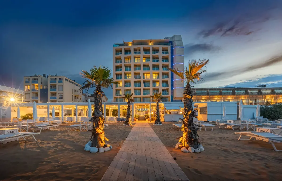 Beachside hotel with palm trees, lounge chairs, and a central walkway, illuminated at dusk.