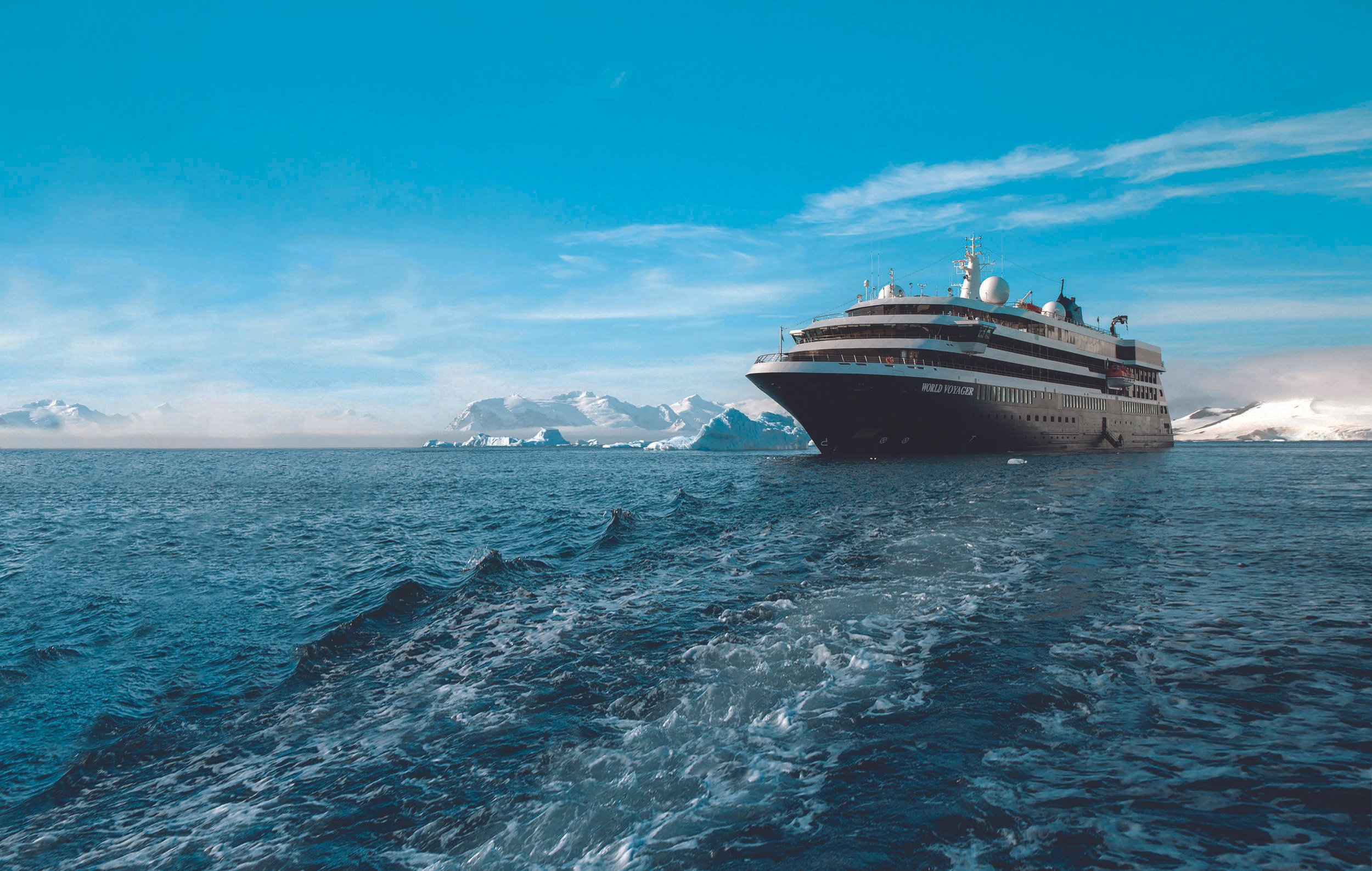 A large cruise ship named 'World Voyager' sailing in icy waters with icebergs and snow-covered mountains in the background under a clear blue sky.