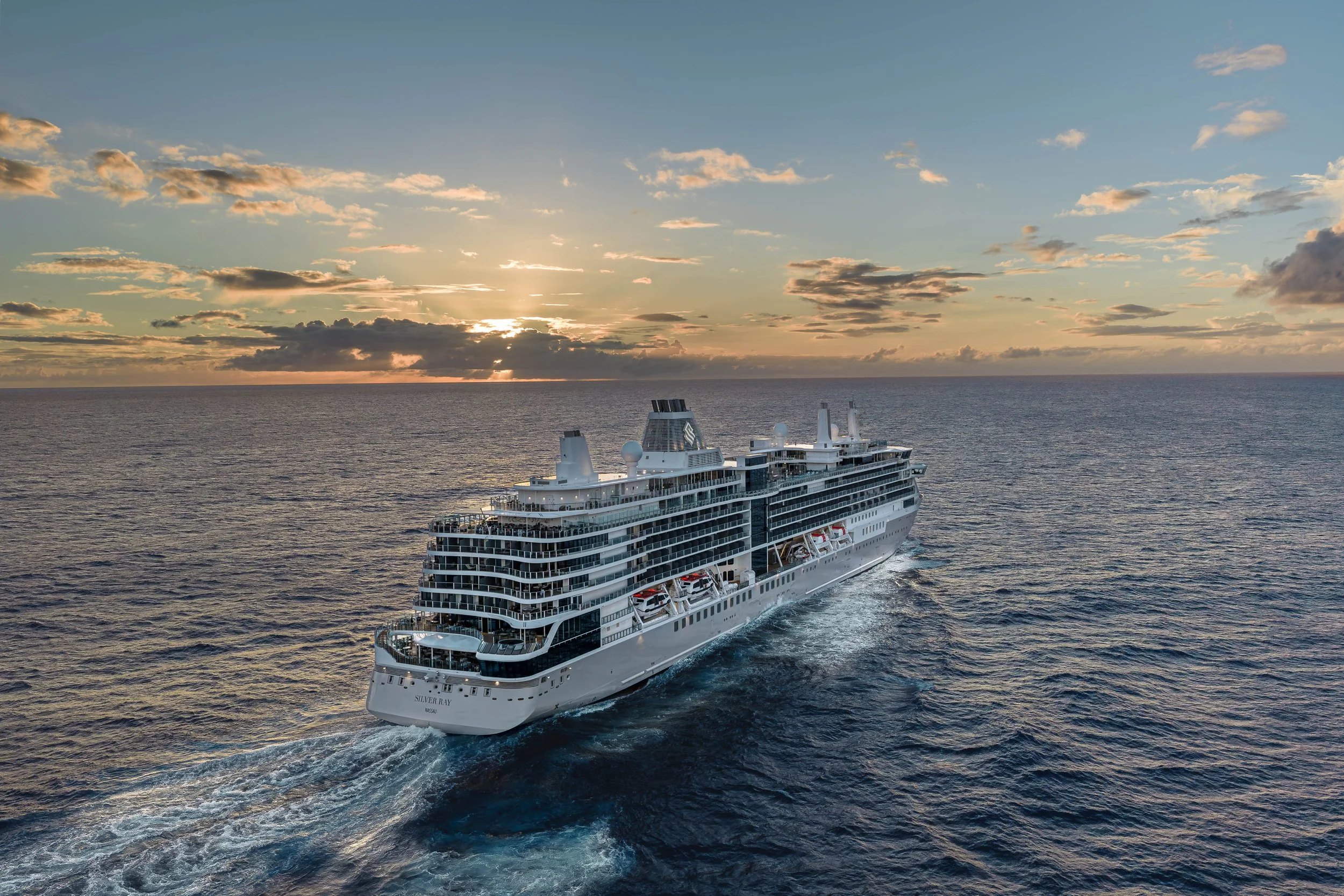 A large white cruise ship sailing on the ocean during sunset with a partly cloudy sky.