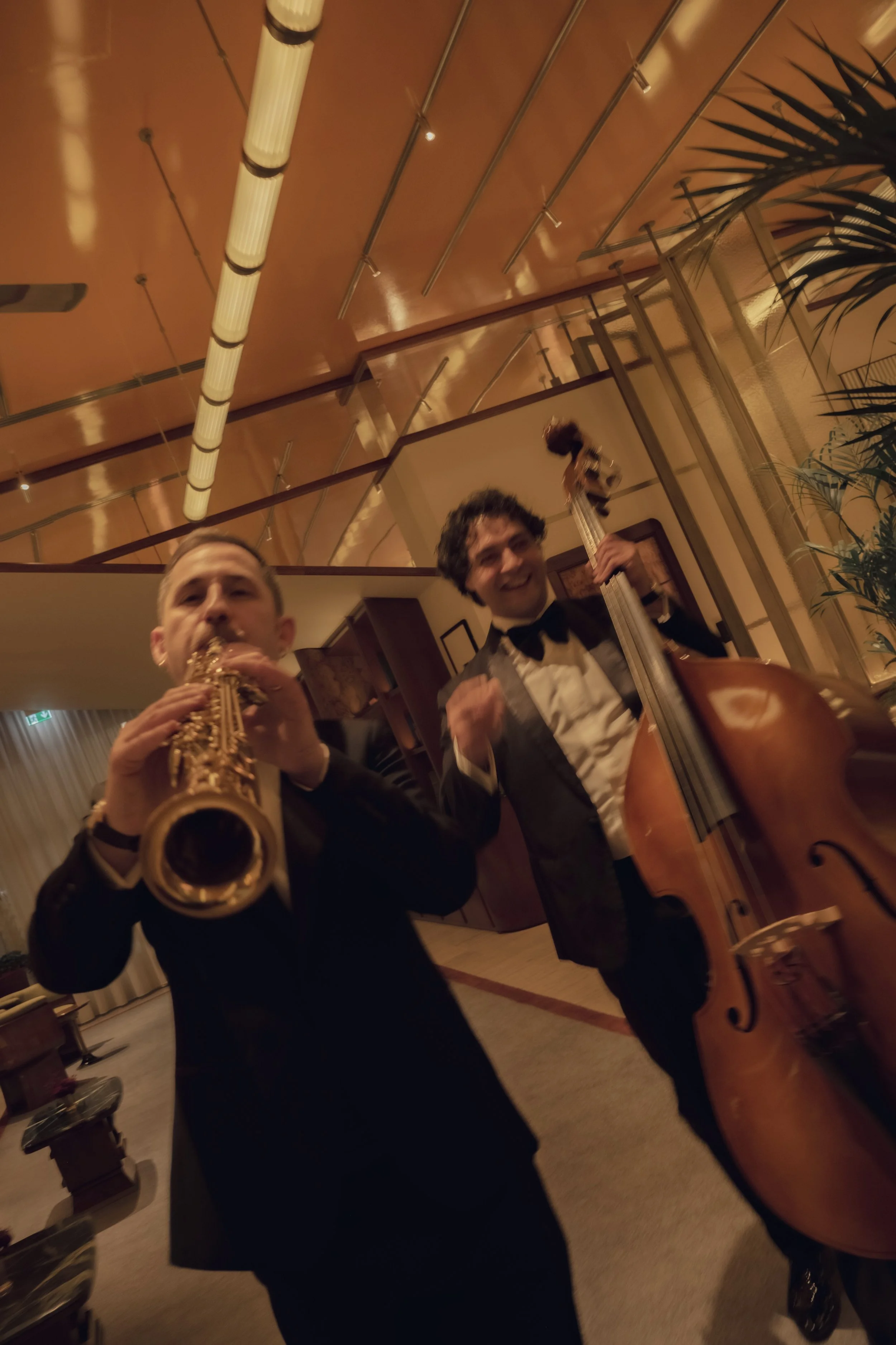 Two men in tuxedos playing musical instruments, one with a saxophone and the other with a double bass, in a warm-lit indoor setting with a ceiling featuring exposed modular panels and a potted plant on the right side.