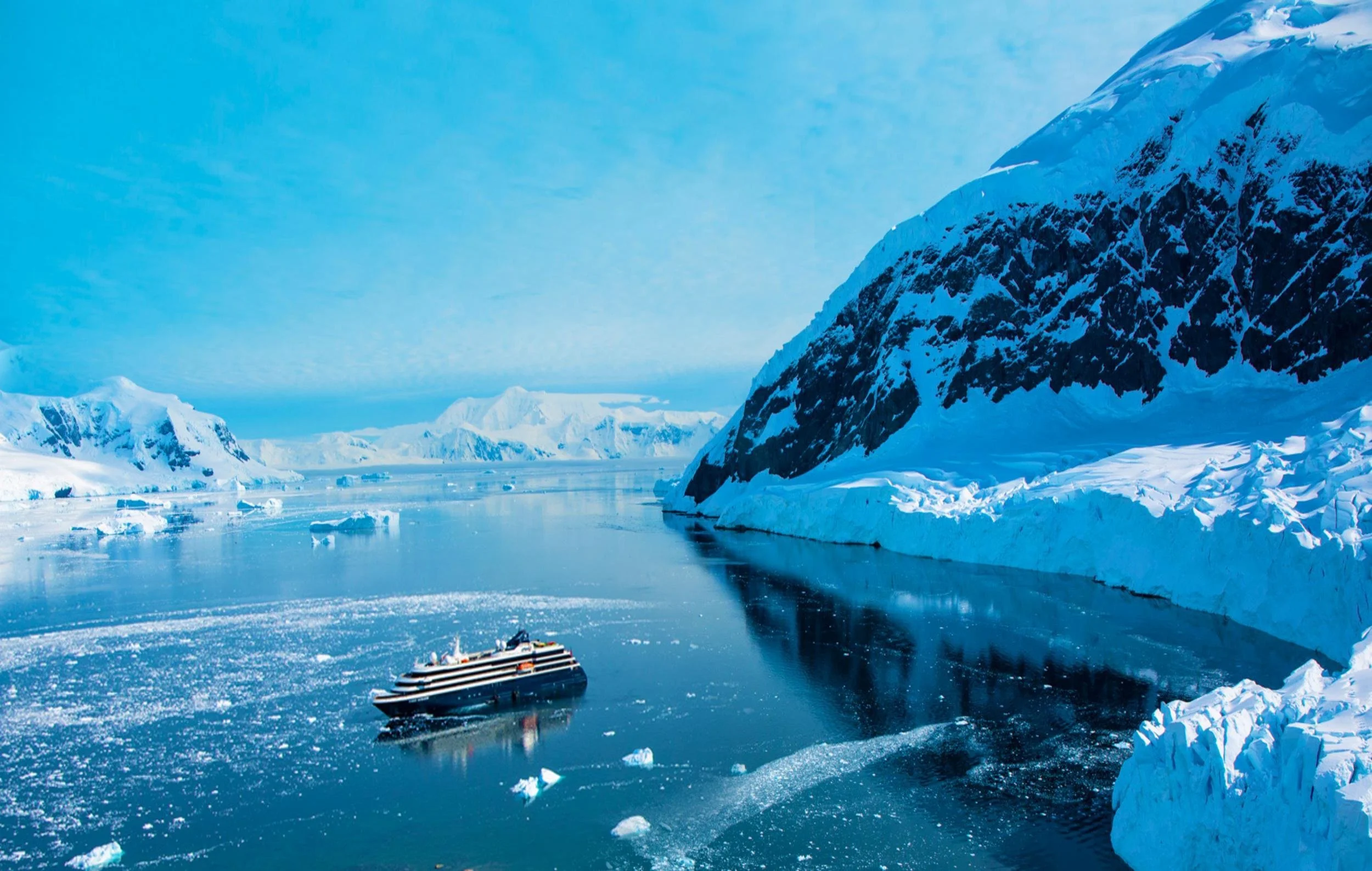 A cruise ship navigating icy waters surrounded by snow-covered mountains and icebergs in a polar region.