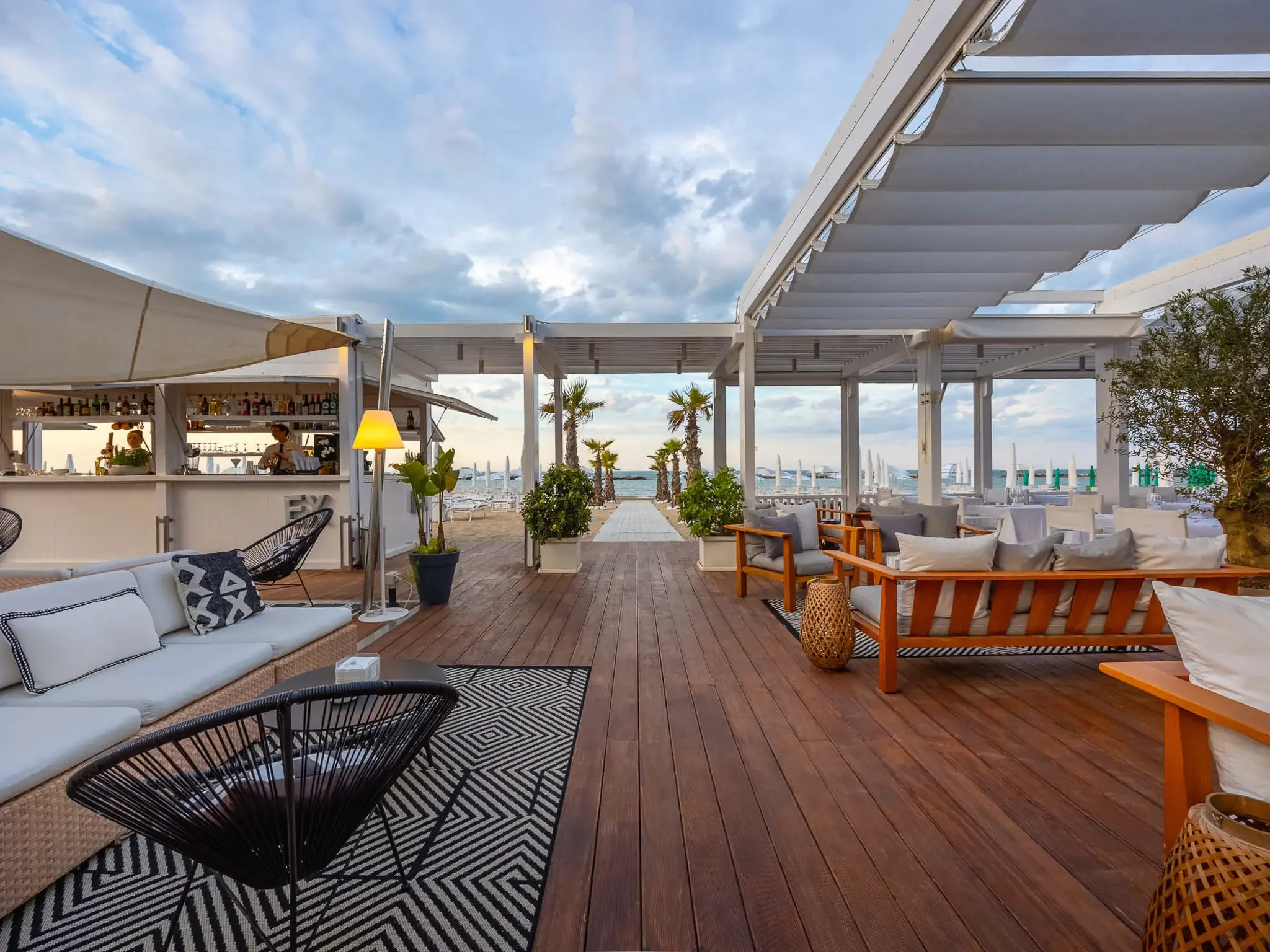 Outdoor coastal restaurant with white pergolas, palm trees, and ocean view, featuring seating areas with sofas, chairs, and tables.