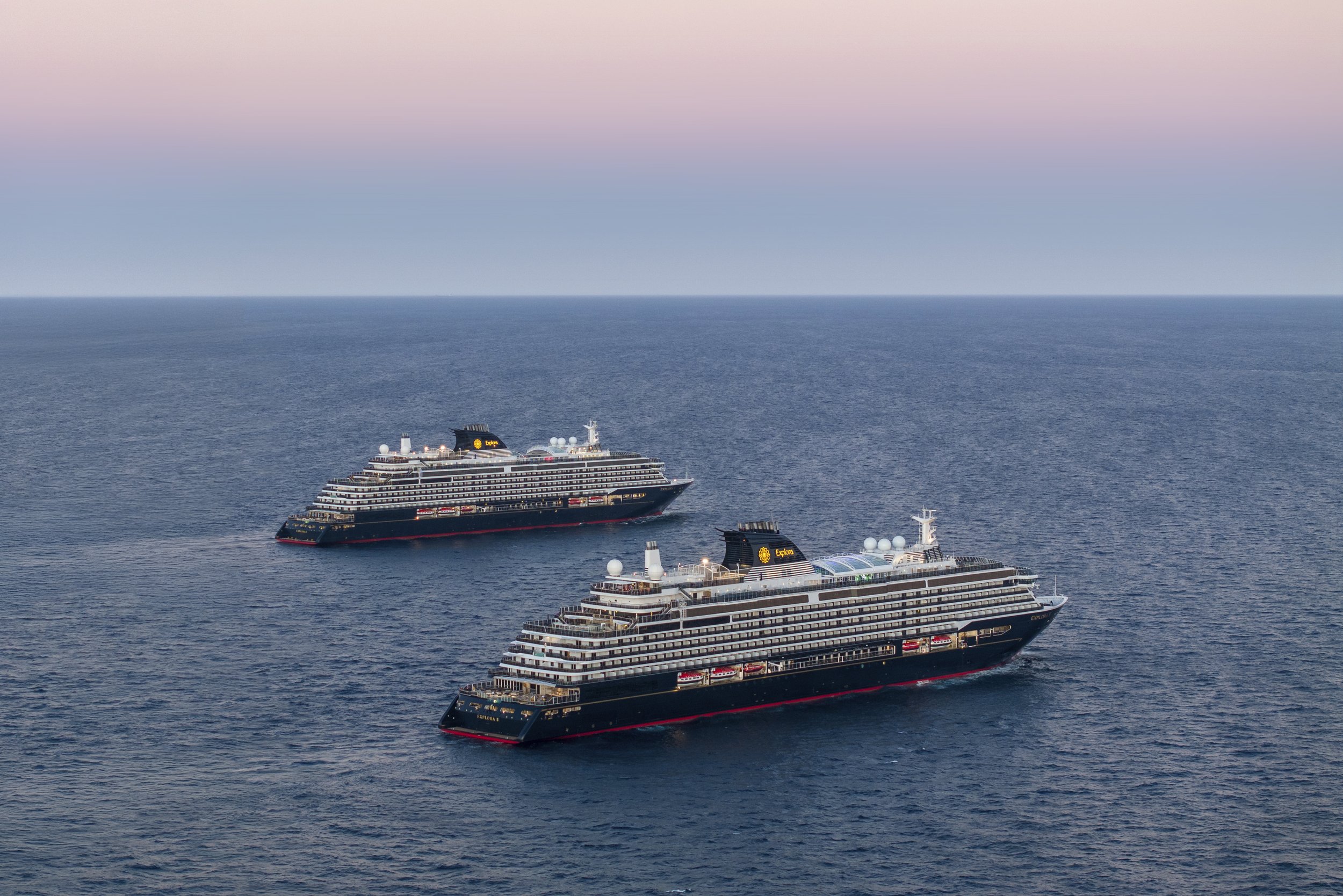 Several large cruise ships sailing on calm ocean waters during sunset or sunrise, with a color gradient in the sky from pink to blue.