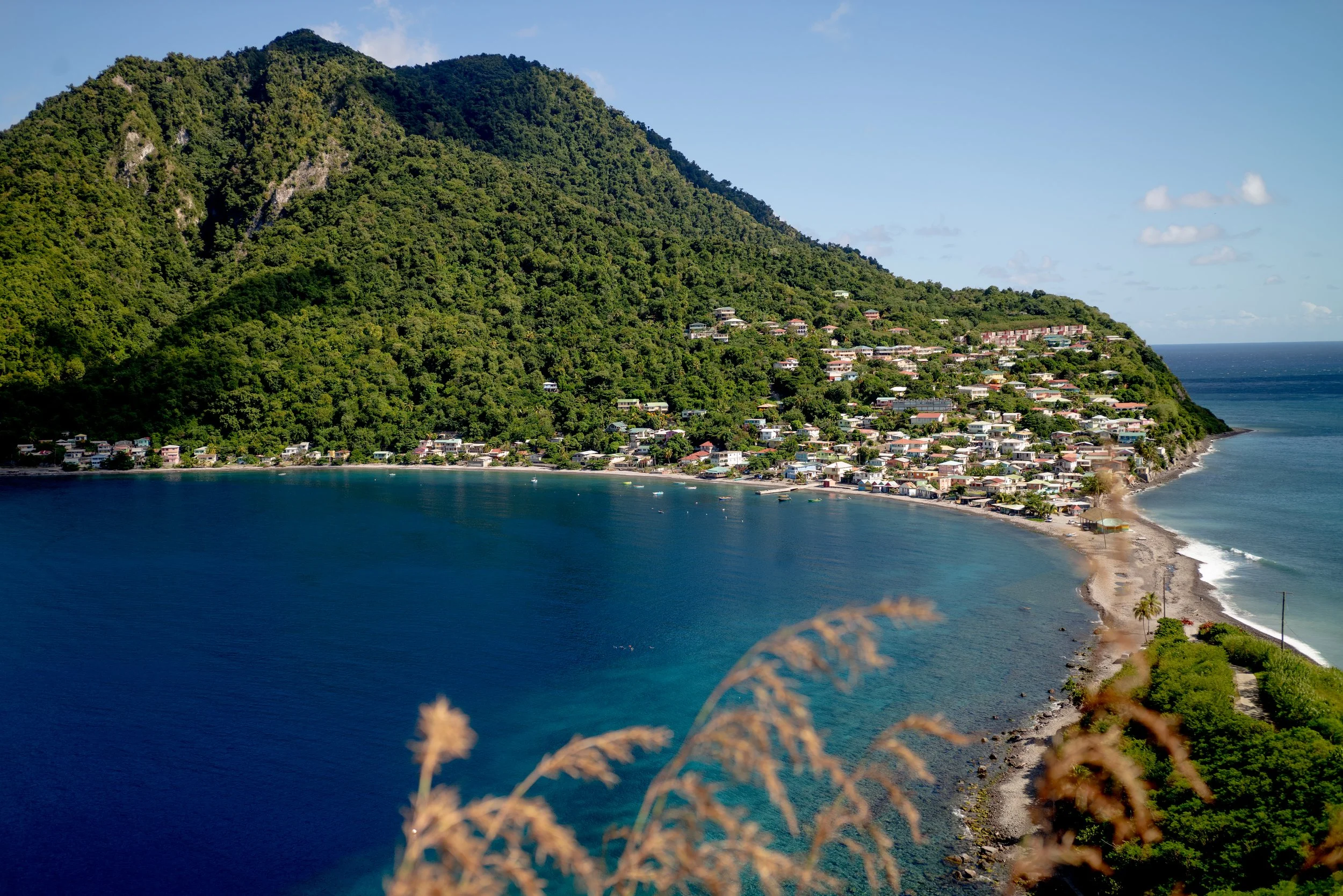 Scenic view of a coastal hillside town with houses nestled on a lush green mountain and a sandy beach along the ocean.