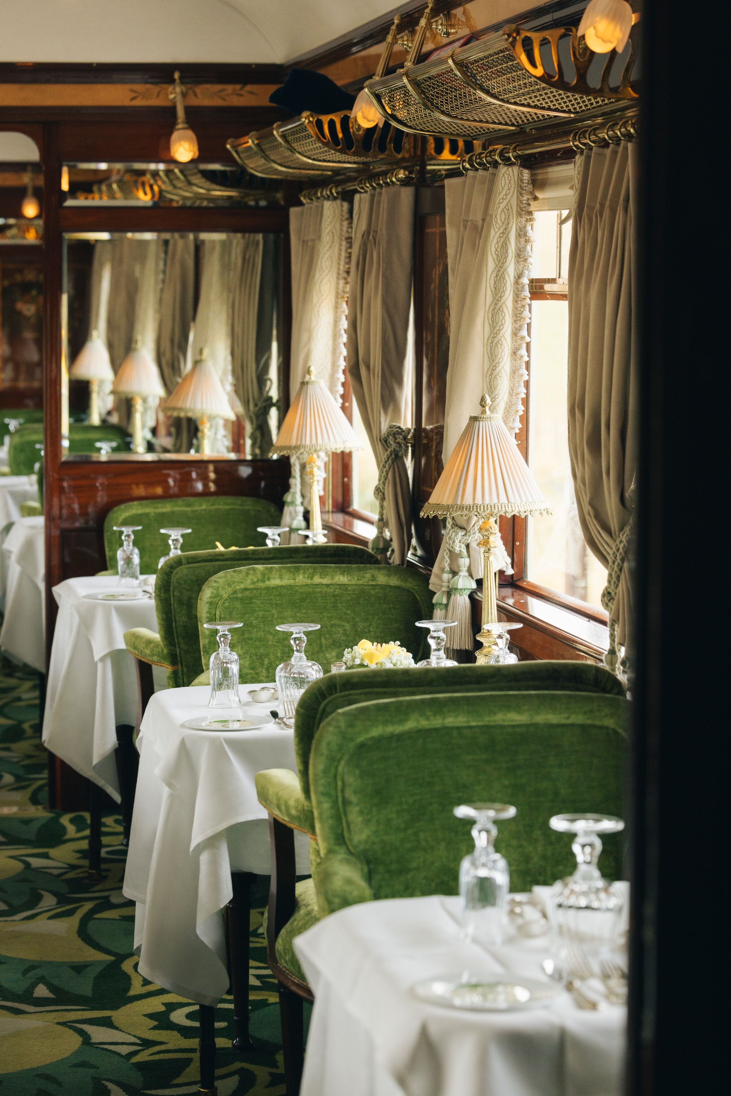 Elegant restaurant interior with white tablecloths, green velvet chairs, table lamps, and windows with curtains, set for dining.