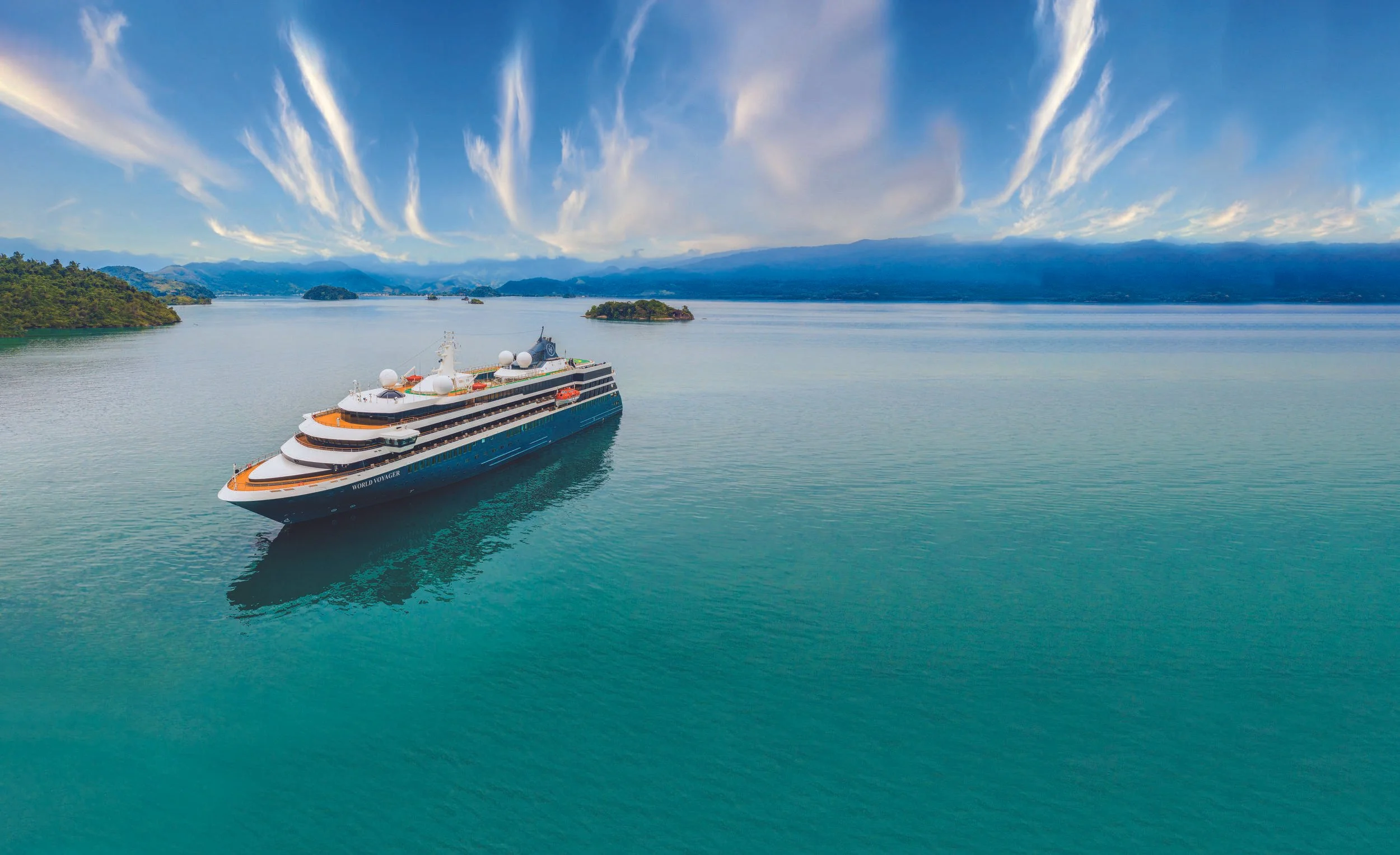 A large cruise ship sailing on a calm body of water with lush green islands and distant mountains under a partly cloudy sky.