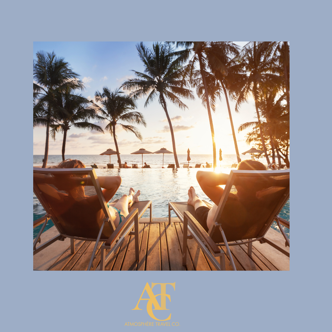 Two people relaxing in lounge chairs by a pool, watching the sunset over a tropical beach with palm trees and umbrellas.