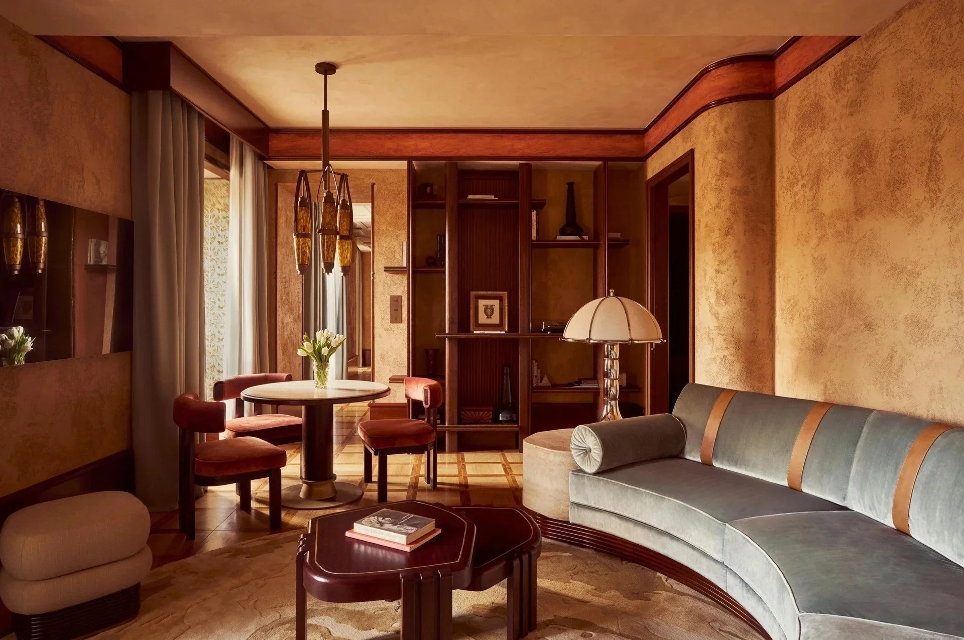 Living room with beige textured walls, wooden accents, a gray curved sofa with a cylinder pillow, and a wooden coffee table with books. There is a round dining table with red upholstered chairs, flowers, and a window with light-colored curtains. A wooden shelf with decorative items and framed art is visible, along with a hanging light fixture and a table lamp.
