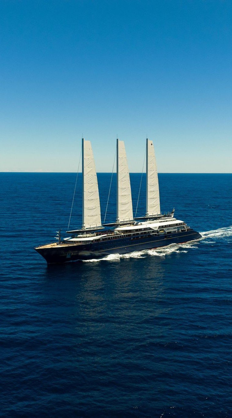 A large luxury sailing yacht with three tall masts sailing on the open ocean under a clear blue sky.