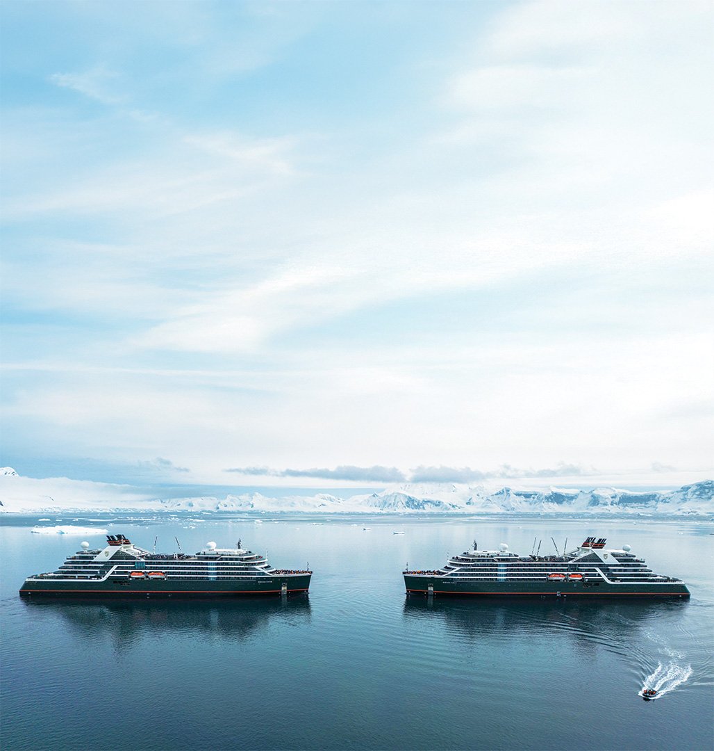 Two large cruise ships sailing in icy Arctic waters with snow-covered mountains and icebergs in the background, under a partly cloudy sky.