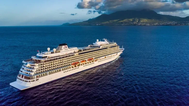 A large white cruise ship sailing in the ocean near a mountain with cloudy sky overhead.