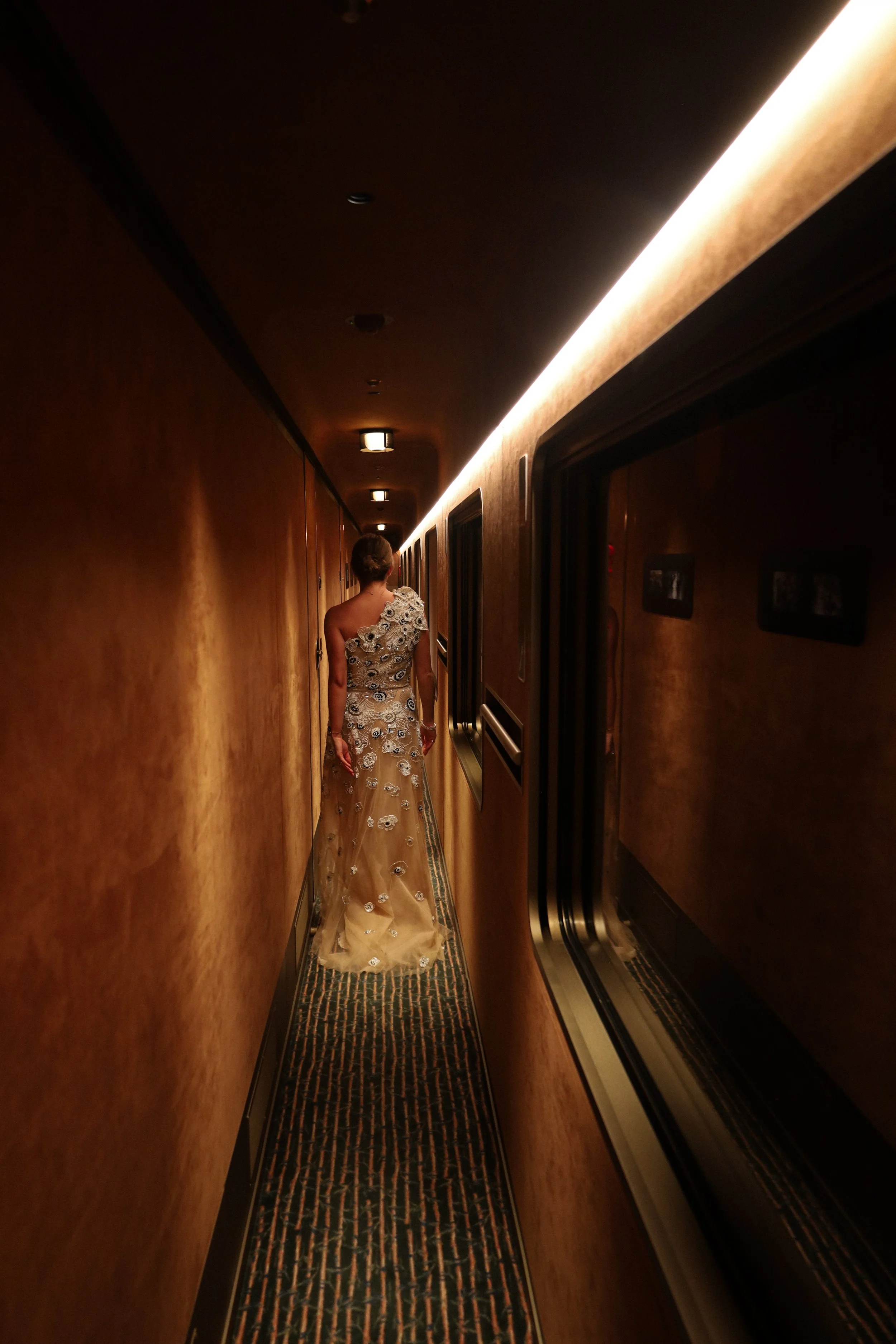 Woman in an elegant dress walking down a narrow corridor with warm lighting on the ceiling.