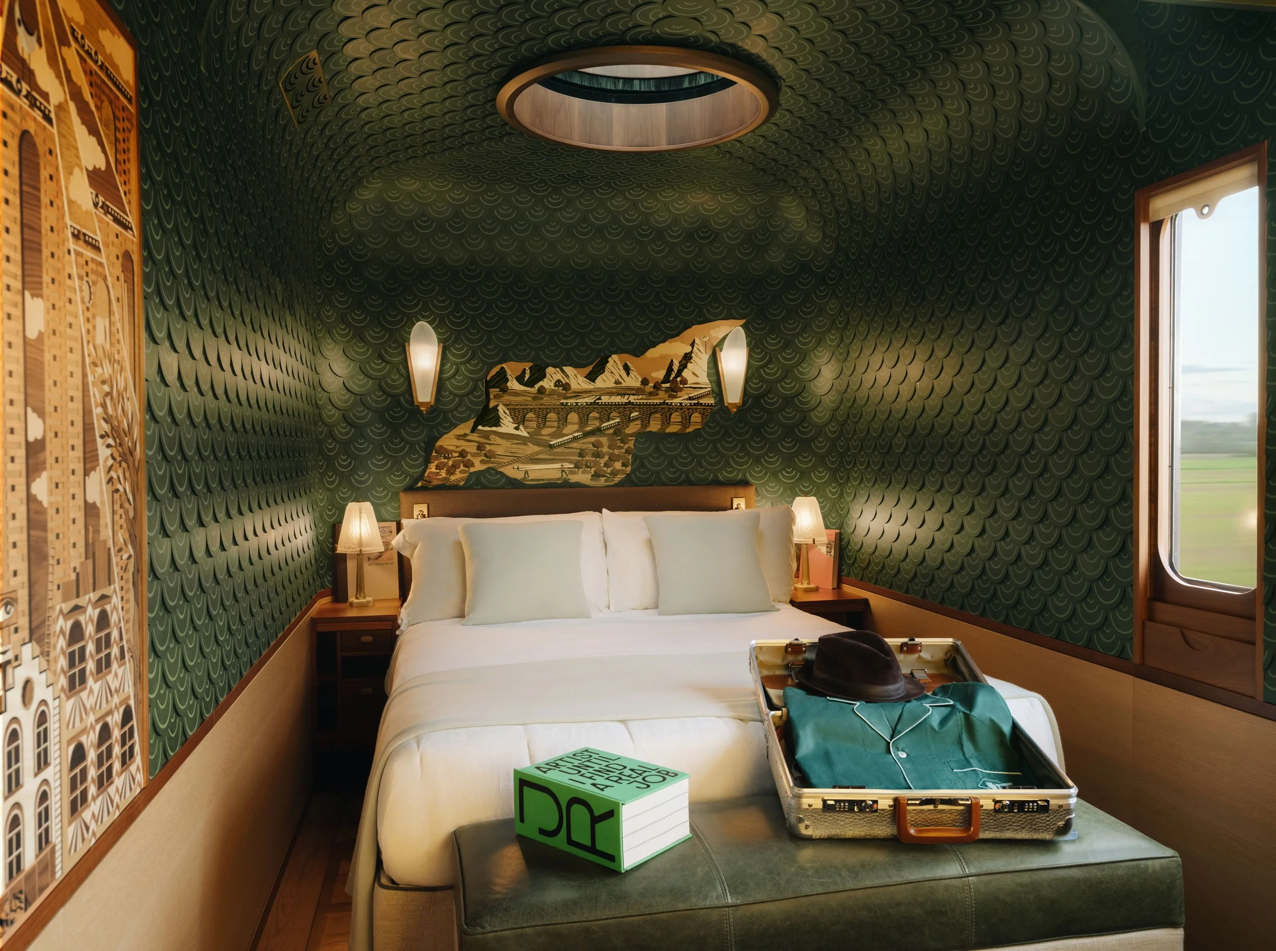 A hotel room with green textured wallpaper, a window with a view outside, a bed with white bedding, a bedside table with lamps, and an open suitcase on the bed containing clothes. There is also a green and white box labeled "Artist Where" on the bed near the suitcase.