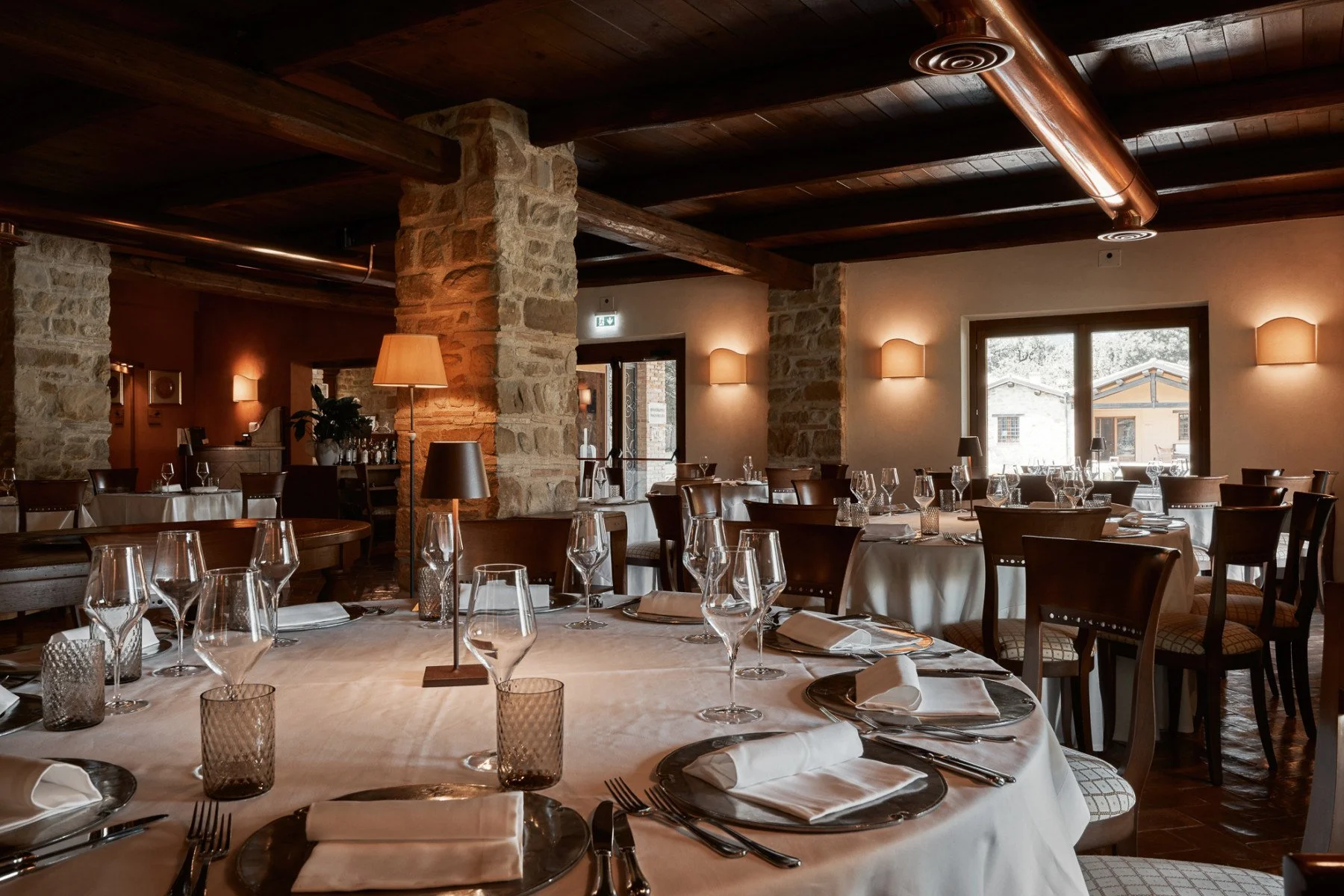 Elegant restaurant interior with round dining tables draped in white tablecloths, set with napkins, wine glasses, and silverware. Warm lighting, stone columns, wooden beams, and large windows create a cozy ambiance.